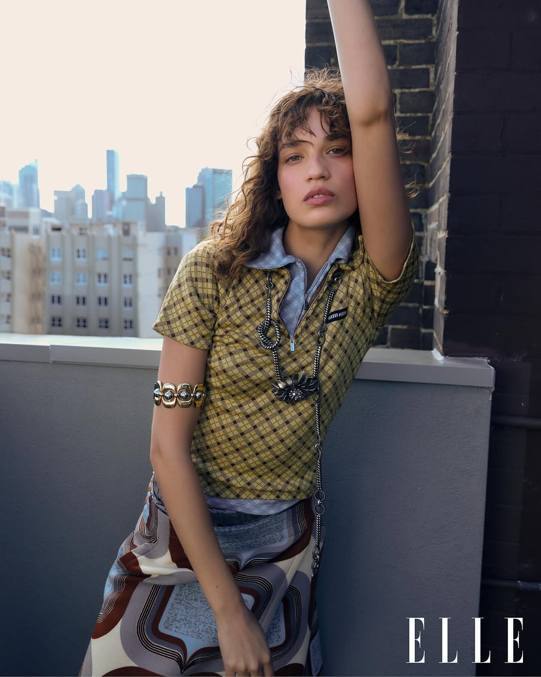 Portrait of a young woman with curly hair in yellow plaid shirt and patterned skirt posing on a rooftop with city buildings in the background.
