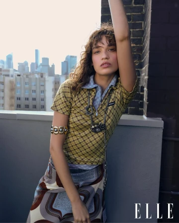 Portrait of a young woman with curly hair in yellow plaid shirt and patterned skirt posing on a rooftop with city buildings in the background.