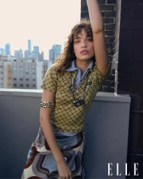 Model wearing a patterned yellow polo top and patterned skirt posing on a rooftop with a city skyline in the background.