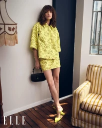 Model wearing yellow patterned set and green mules leaning against an interior wall with a lamp and armchair