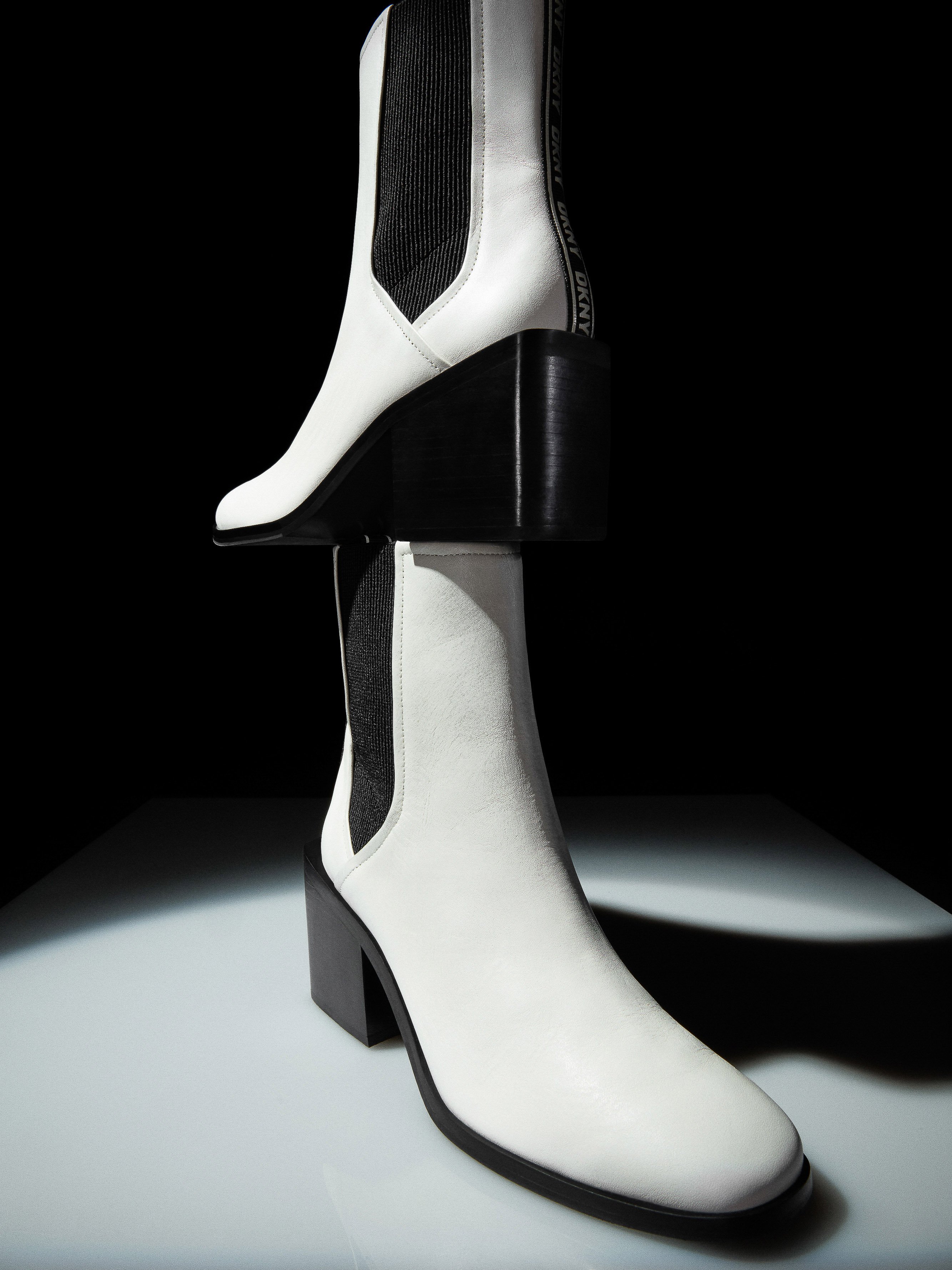 Elevated product shot showcases a pair of white leather heeled ankle boots with black block heels, against a gradient black background.