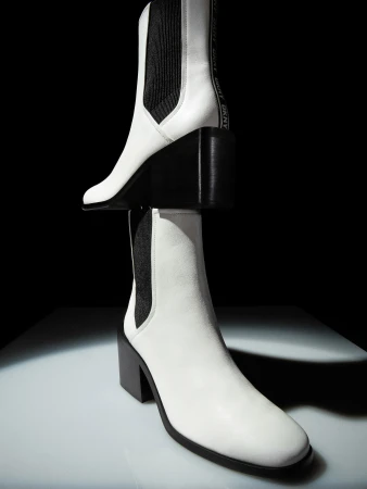 Elevated product shot showcases a pair of white leather heeled ankle boots with black block heels, against a gradient black background.