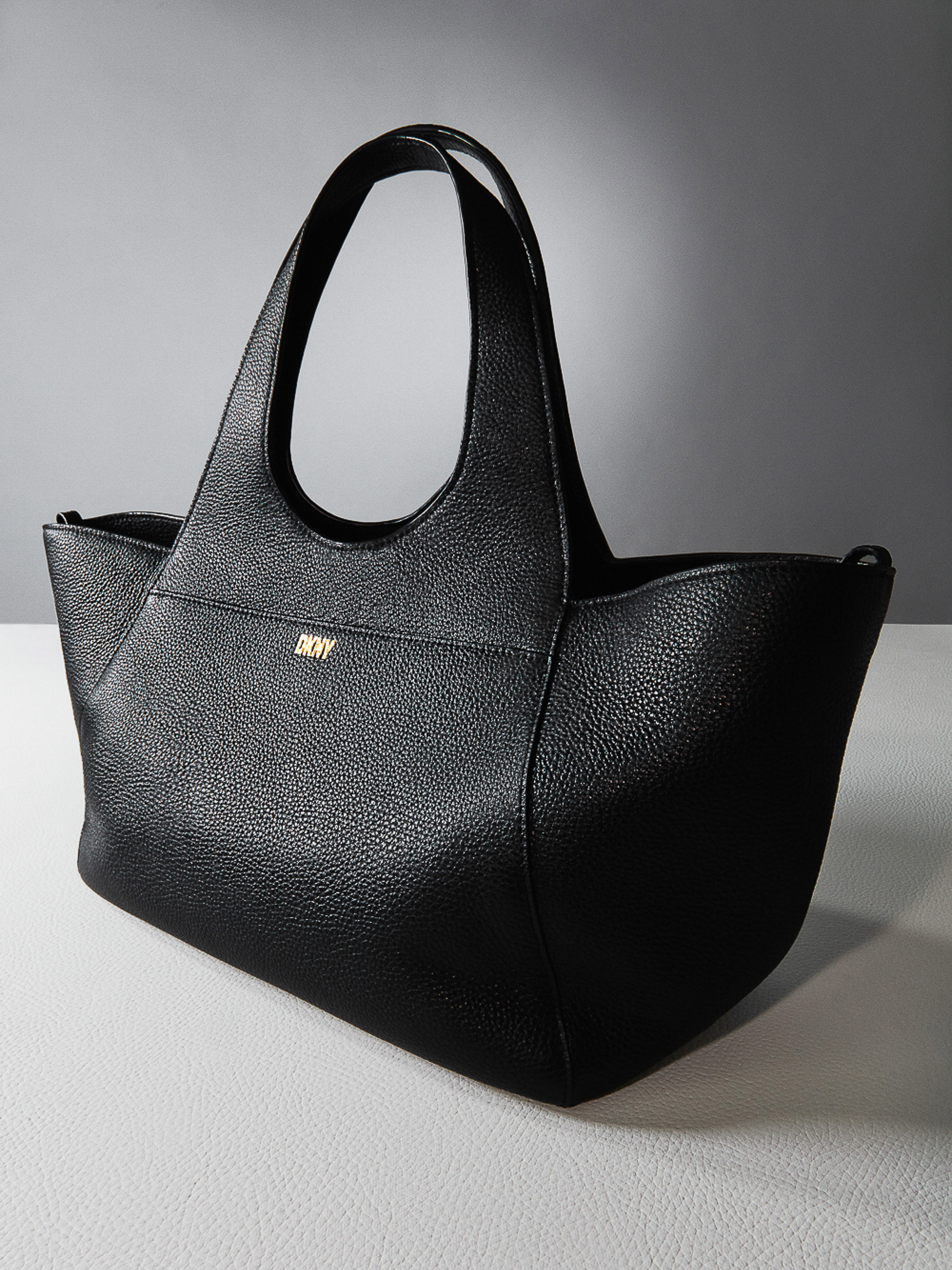 Black DKNY tote bag on a light gray surface against a dark gray background, showcasing its texture and design.