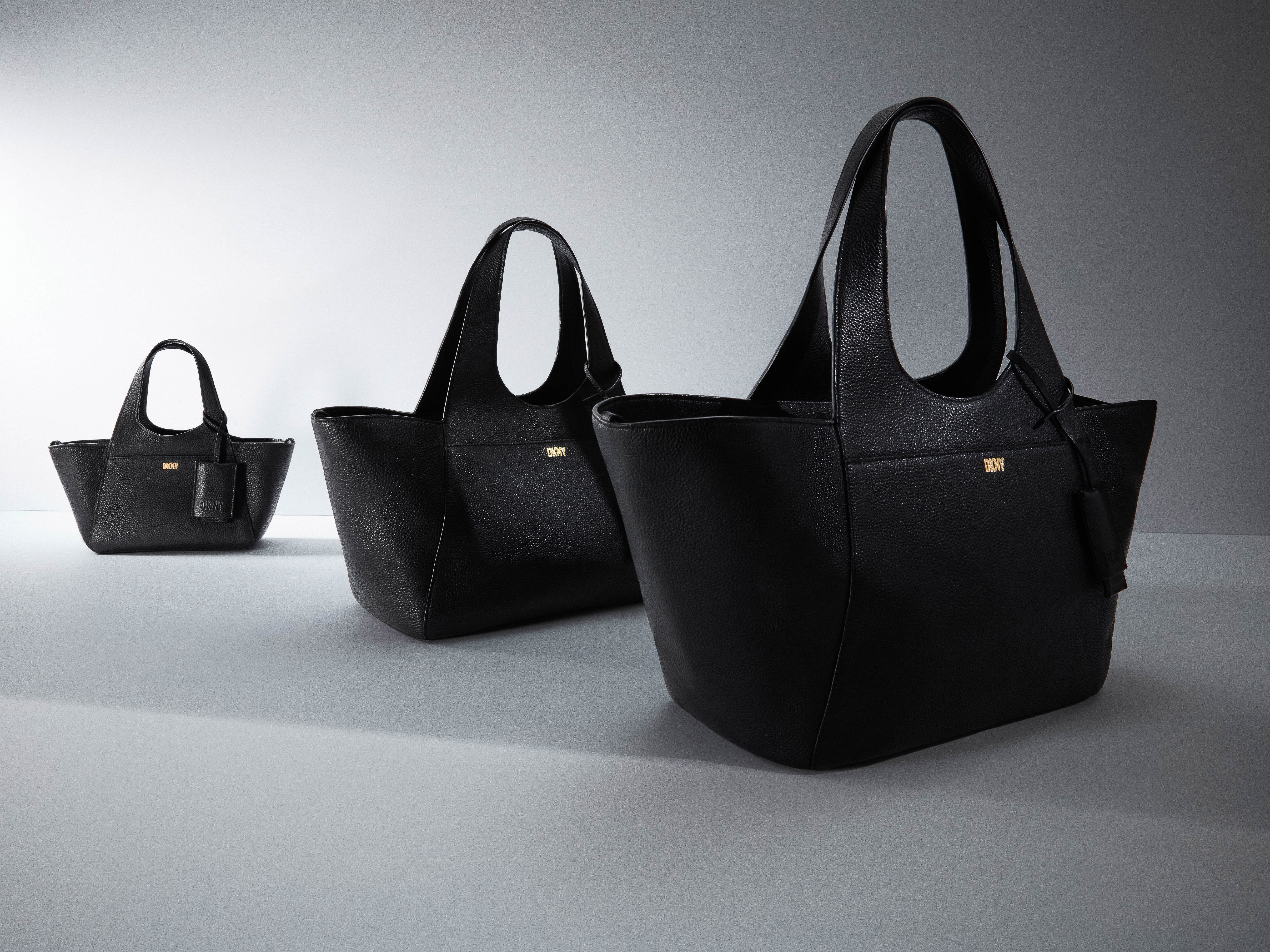 Studio shot of three black textured leather tote bags of varying sizes arranged on a gradient white background.