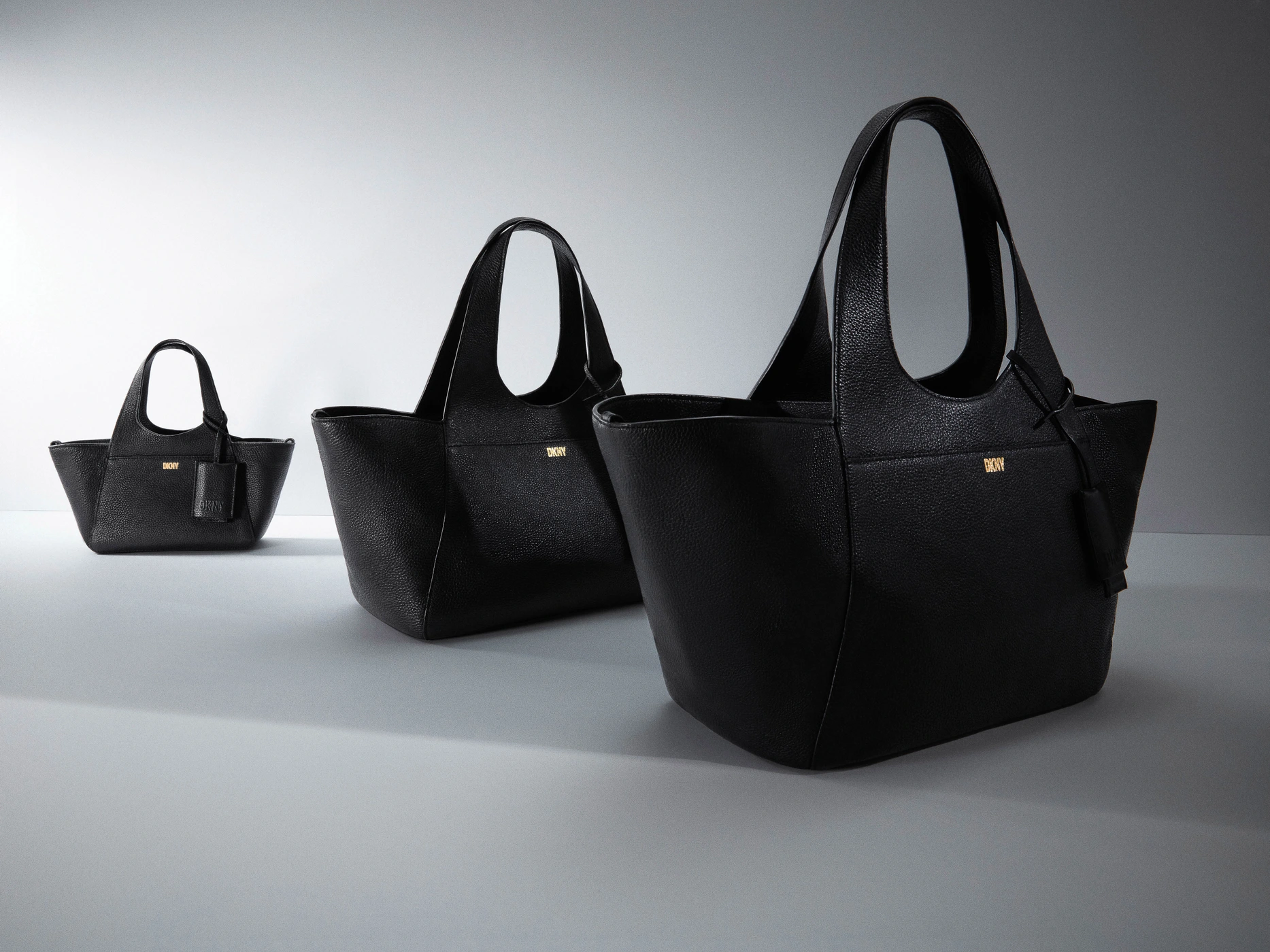 Three black textured leather tote bags of varying sizes arranged diagonally on a light grey studio background.