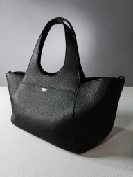Studio product photograph of a black textured leather tote handbag with gold DKNY lettering on a light surface.