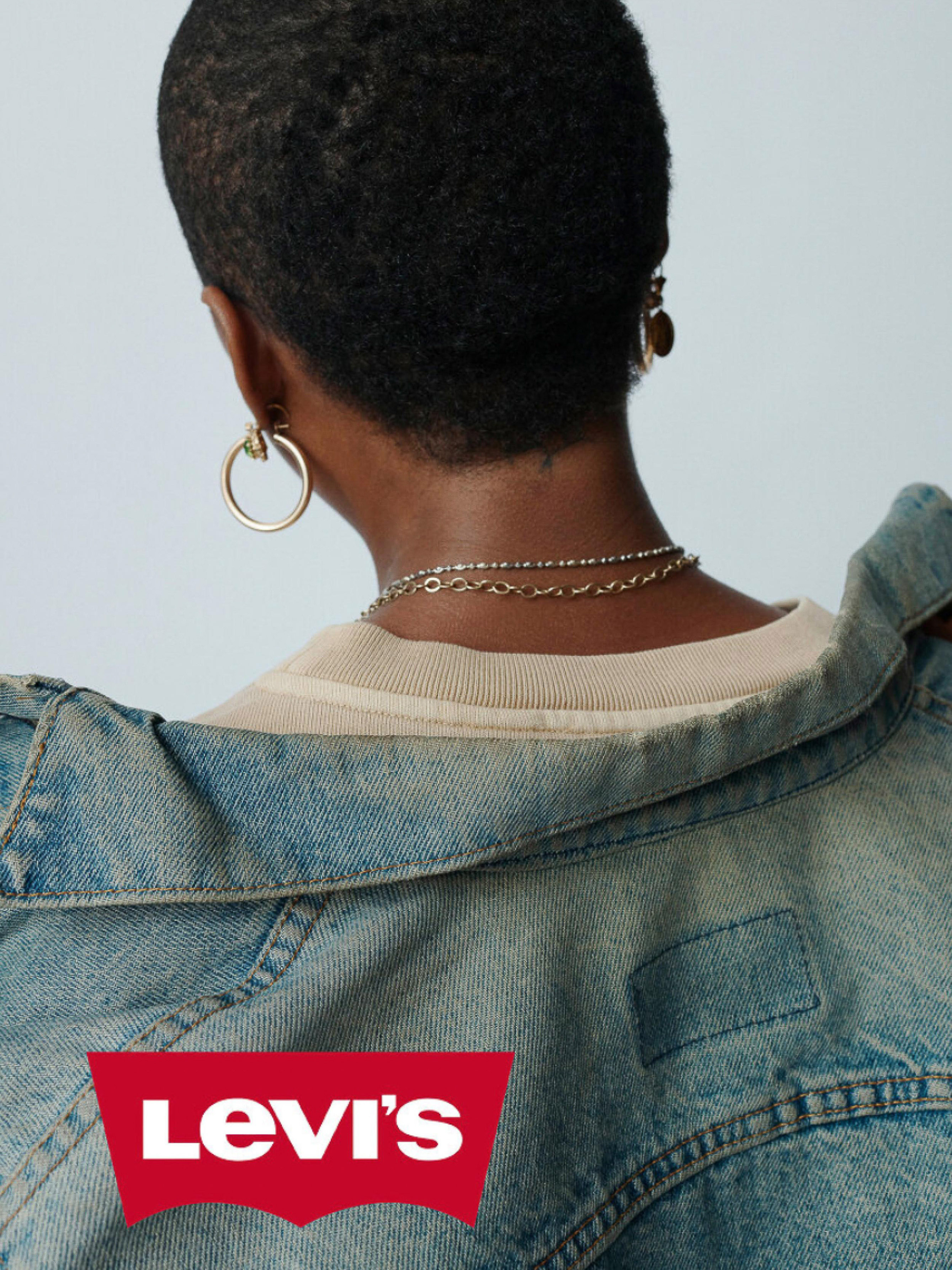 Back view of woman with short dark hair wearing a blue denim jacket over a tan shirt with gold hoop earrings and necklaces.
