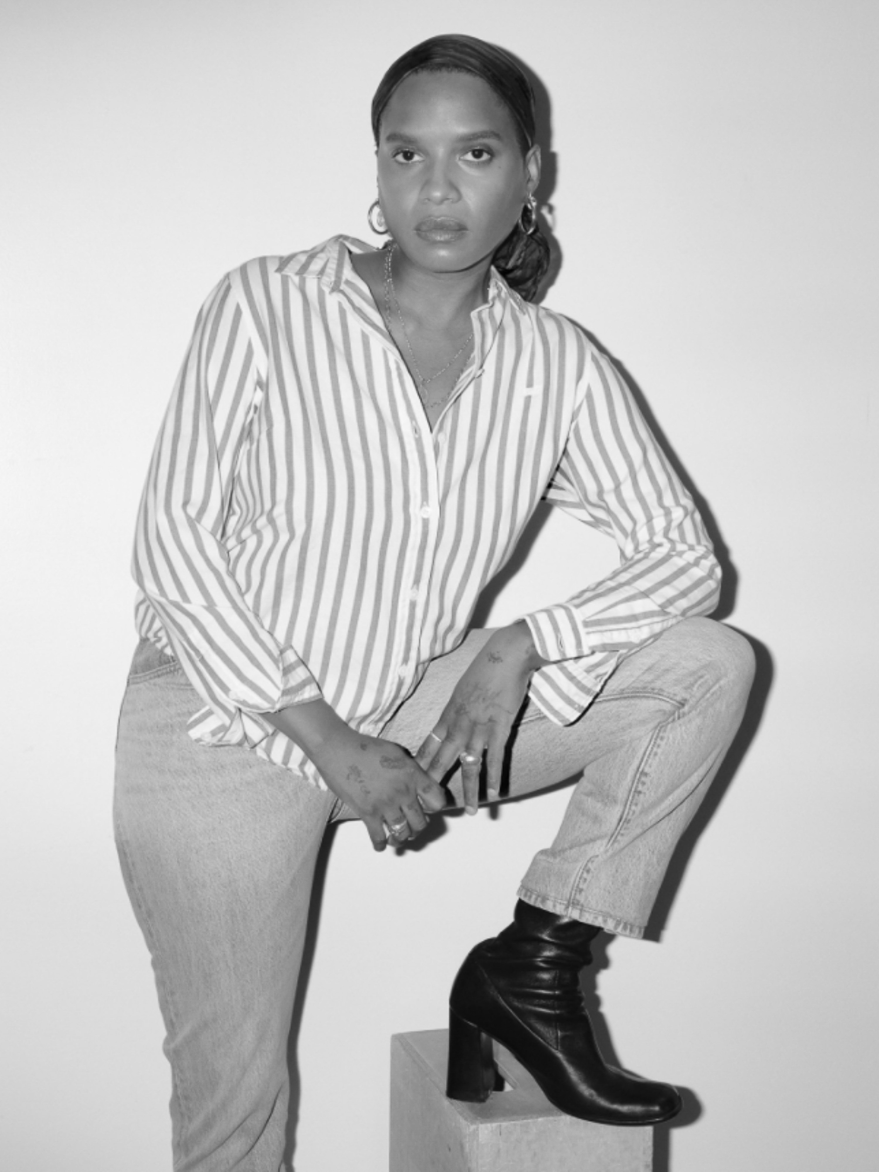 Black and white portrait of a woman posing with a striped shirt, jeans and heeled boots, sitting on a geometric prop against a white wall.