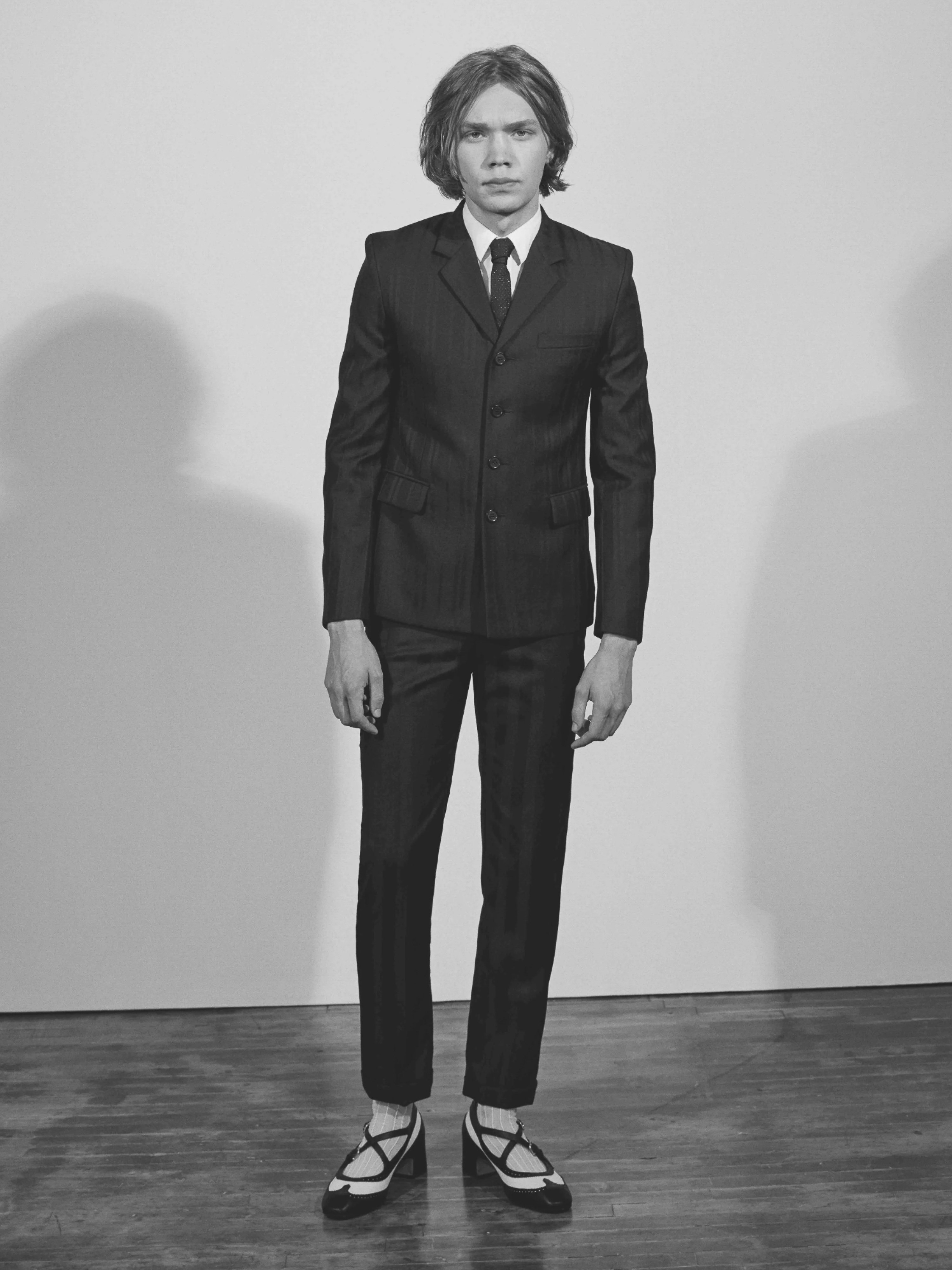 Black and white portrait of a young adult man in a dark pin-striped suit, white shirt, and dark tie standing indoors.