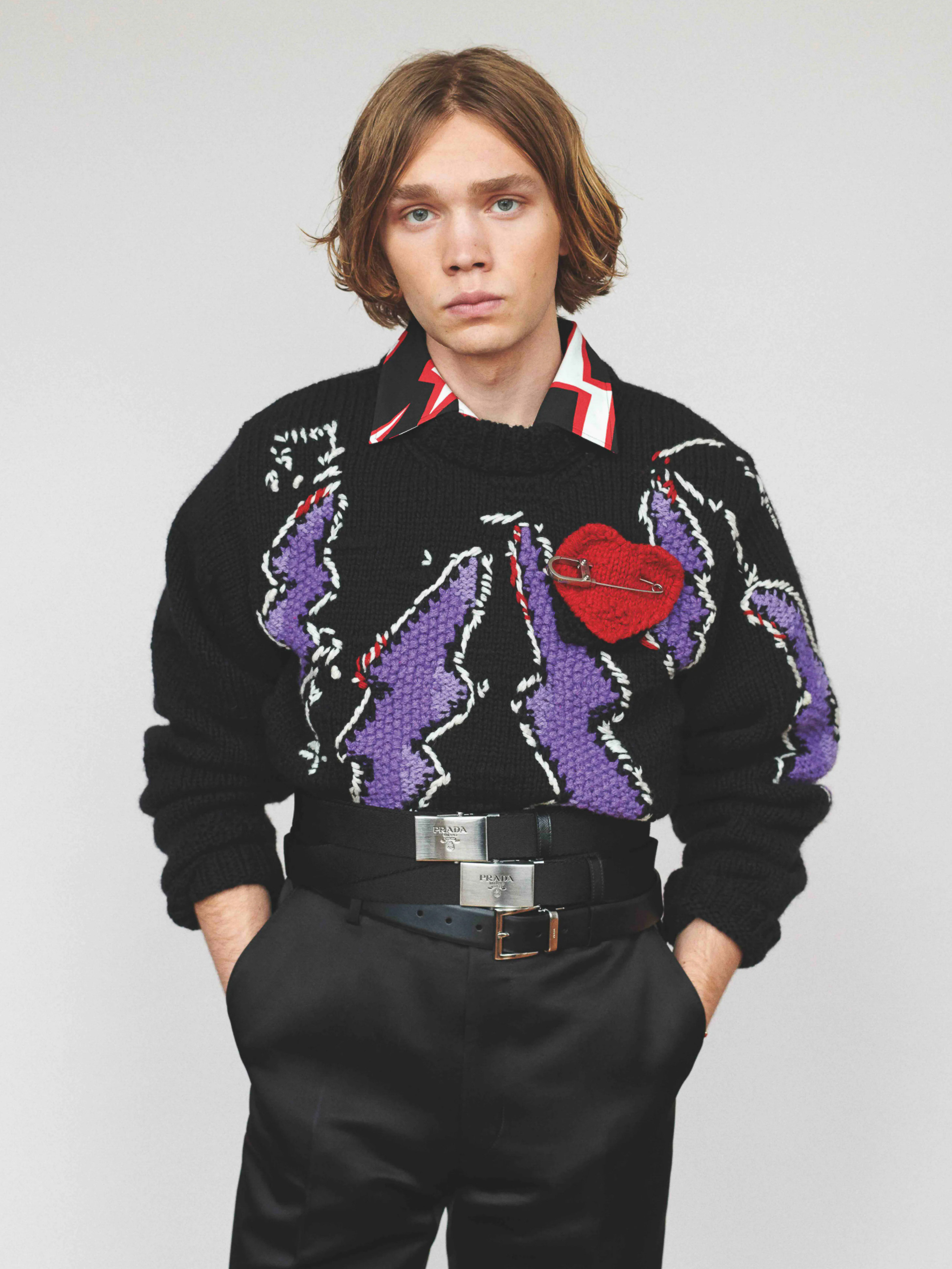 Young man poses wearing a black sweater with purple details, multiple belts, and black pants, set against a plain white backdrop.