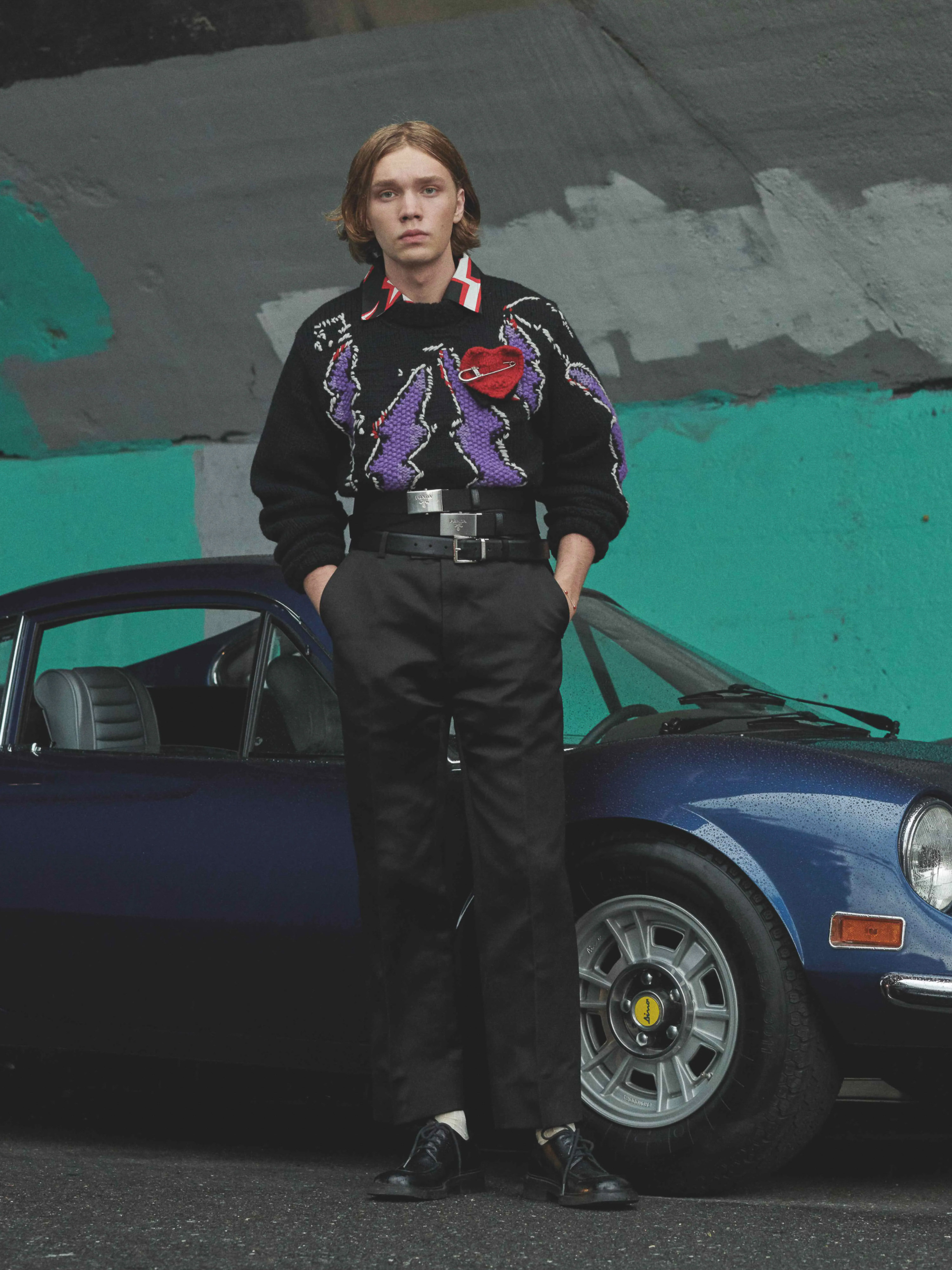 Male fashion model in a sweater standing next to a blue sportscar with an urban concrete background, fashionable and stylish.