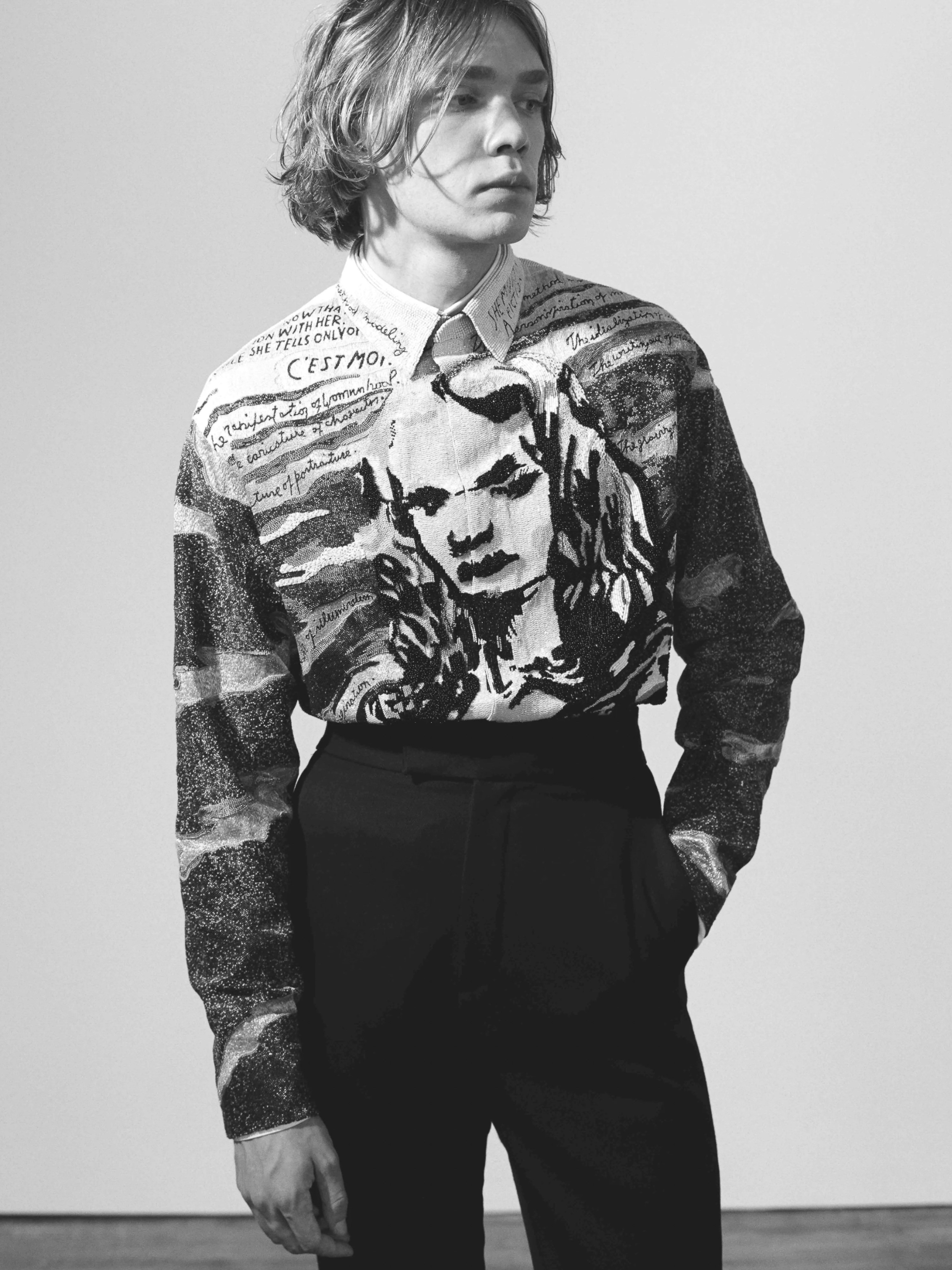 Black and white fashion portrait of a young adult male wearing a printed long-sleeve shirt and dark trousers against a plain wall.