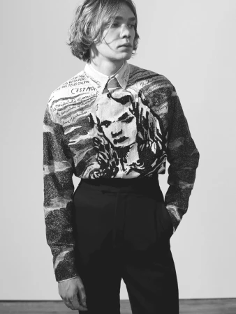 Black and white fashion portrait of a young adult male wearing a printed long-sleeve shirt and dark trousers against a plain wall.