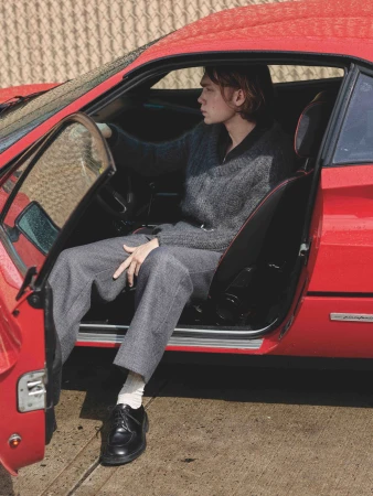 Adult man sits in a red sports car with the door open wearing grey pants, white socks, black shoes, and a grey sweater.