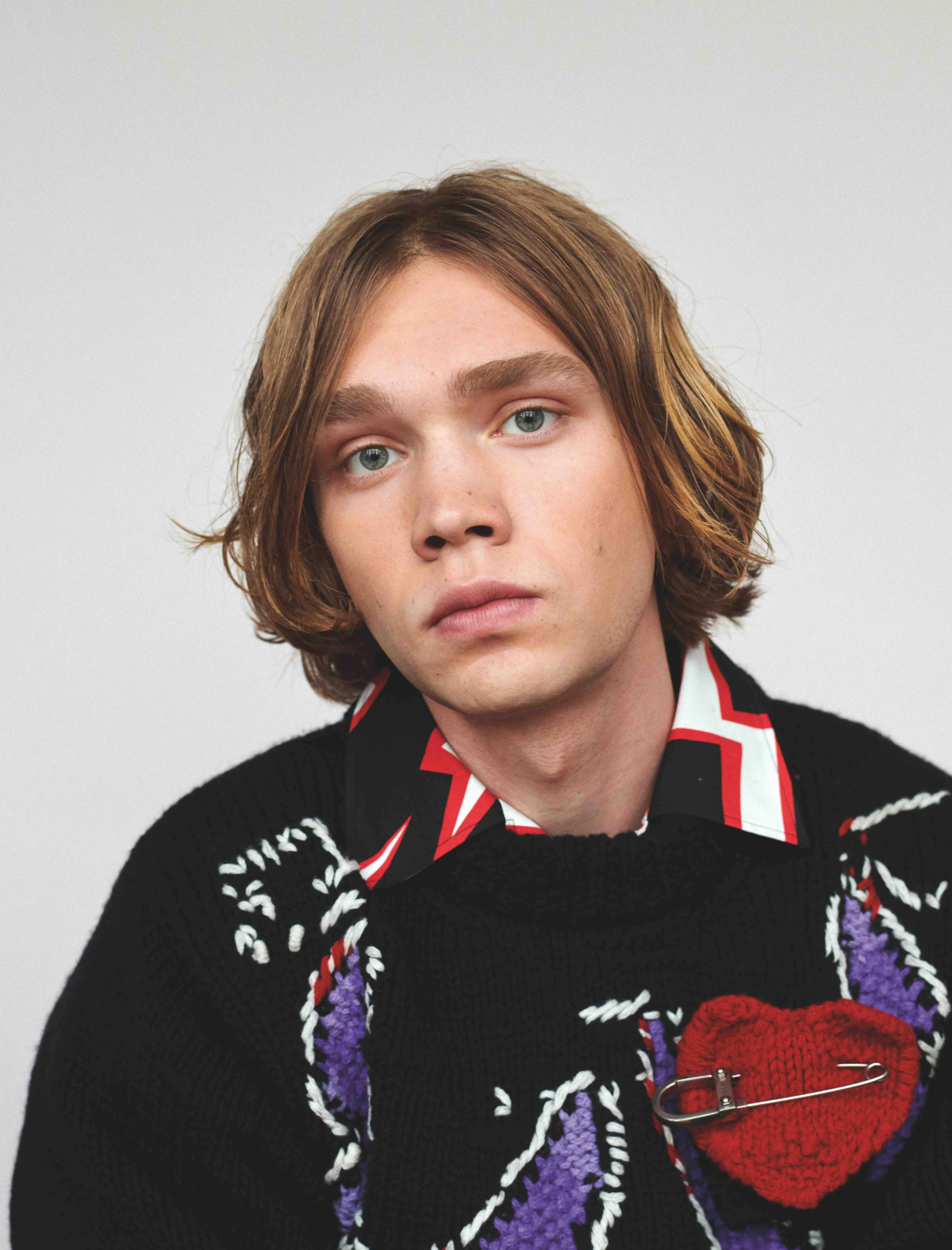 Portrait of a young adult male with blonde hair wearing a black knit sweater with a red heart against a plain white background.