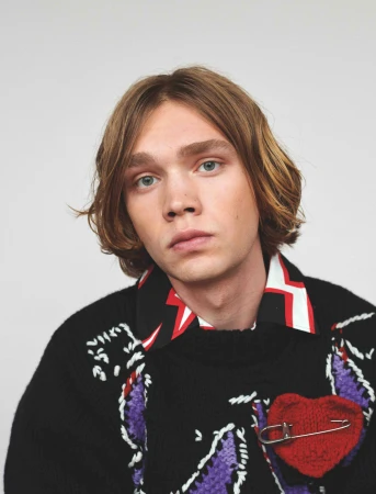 Portrait of a young adult male with blonde hair wearing a black knit sweater with a red heart against a plain white background.