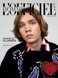 Portrait of actor Charlie Plummer wearing a patterned knit sweater with a heart pin against a plain light background.