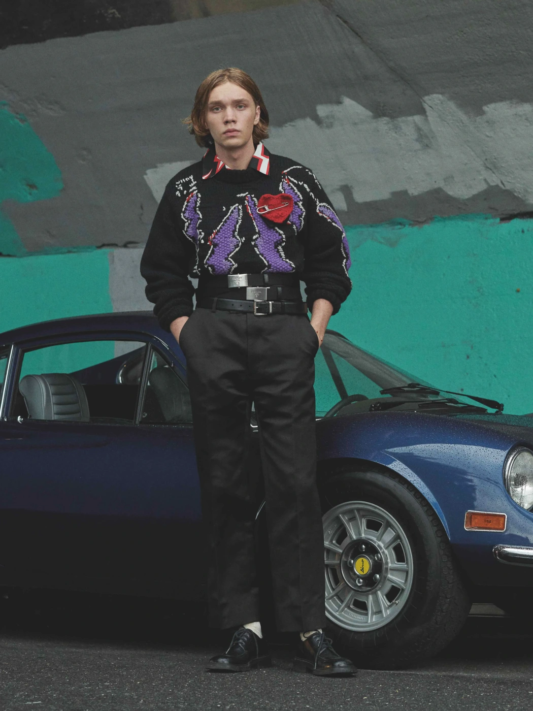 Young man with shoulder-length blonde hair posing in front of a classic blue car against an urban concrete wall.