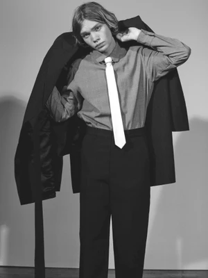 Young man in a button-down shirt and white tie holding a dark blazer over his shoulders against a light studio backdrop