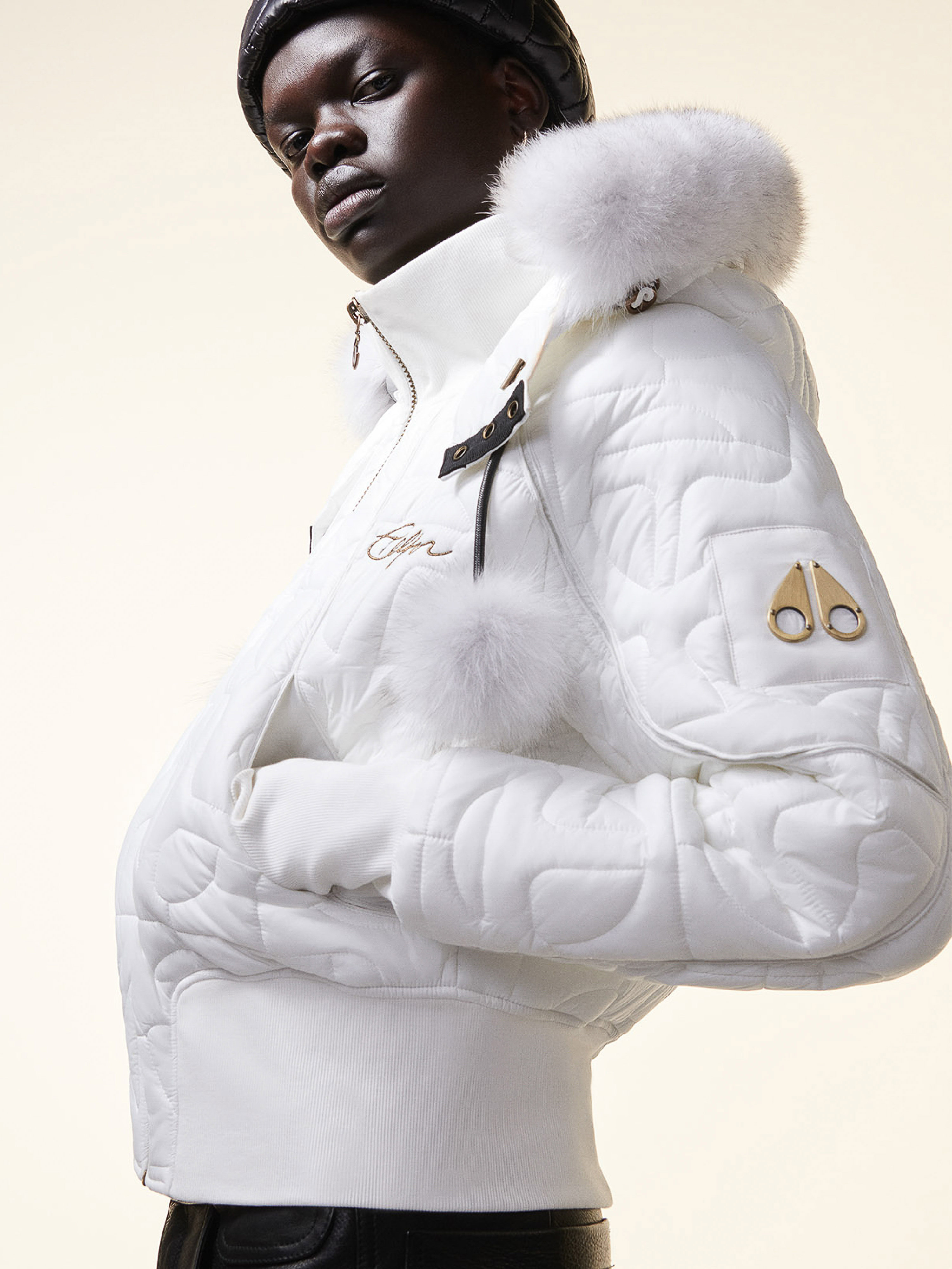 Woman wearing a quilted white jacket with a fur-lined hood poses indoors. The jacket features the brand logo on the sleeve.