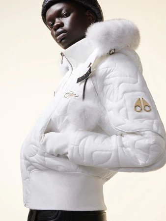Woman wearing a quilted white jacket with a fur-lined hood poses indoors. The jacket features the brand logo on the sleeve.