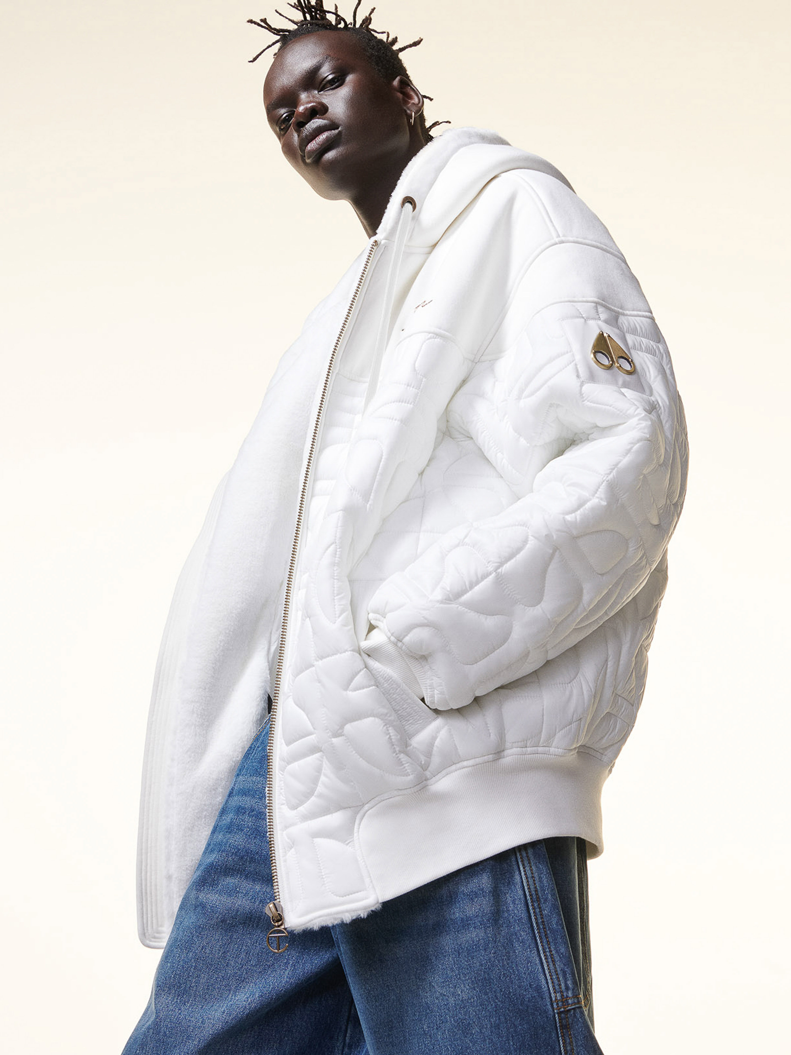 Adult male model posing in a white quilted hooded jacket and blue jeans against a neutral background in a studio fashion shot.