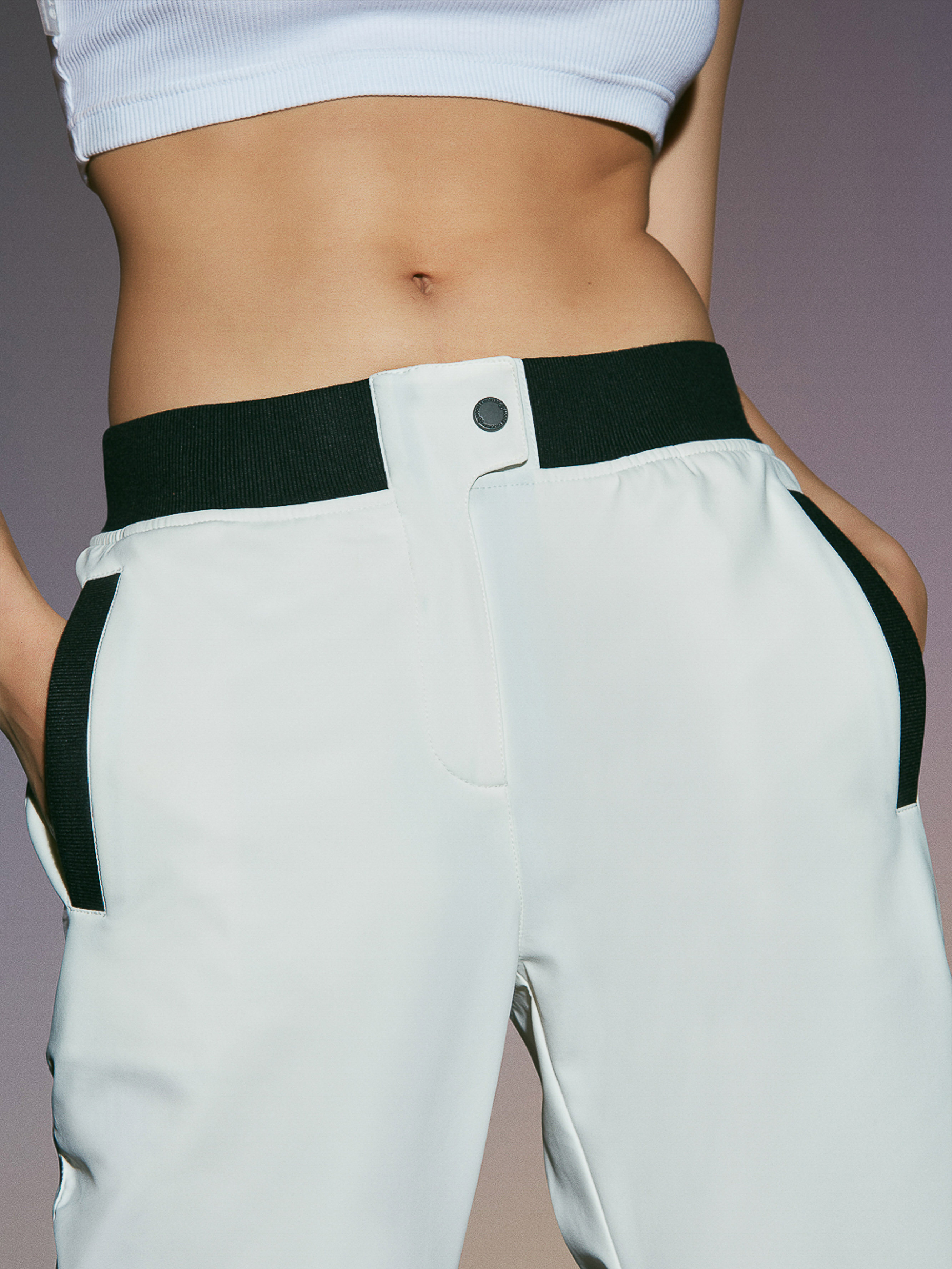 Waist-up shot of a woman wearing a white cropped top and light grey pants with black details against a gradient purple background.