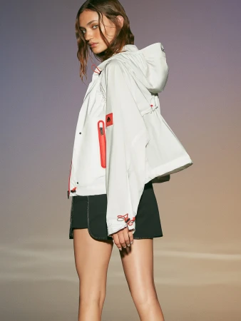 Woman in a hooded white jacket with orange accents and black shorts poses against a soft gradient studio background.