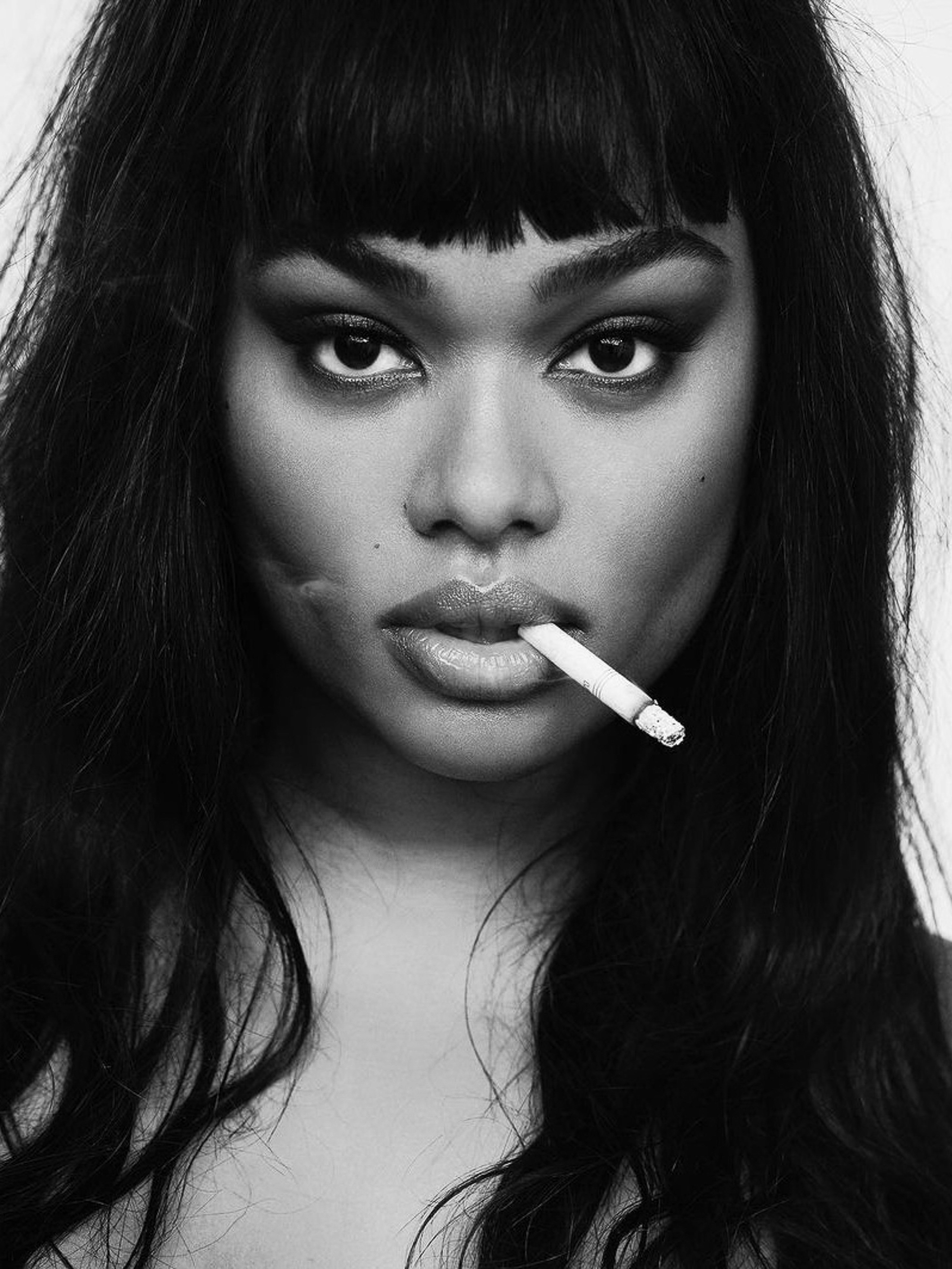 Close-up portrait of an adult woman with dark hair smoking a cigarette, set against a bright background in a monochrome aesthetic.
