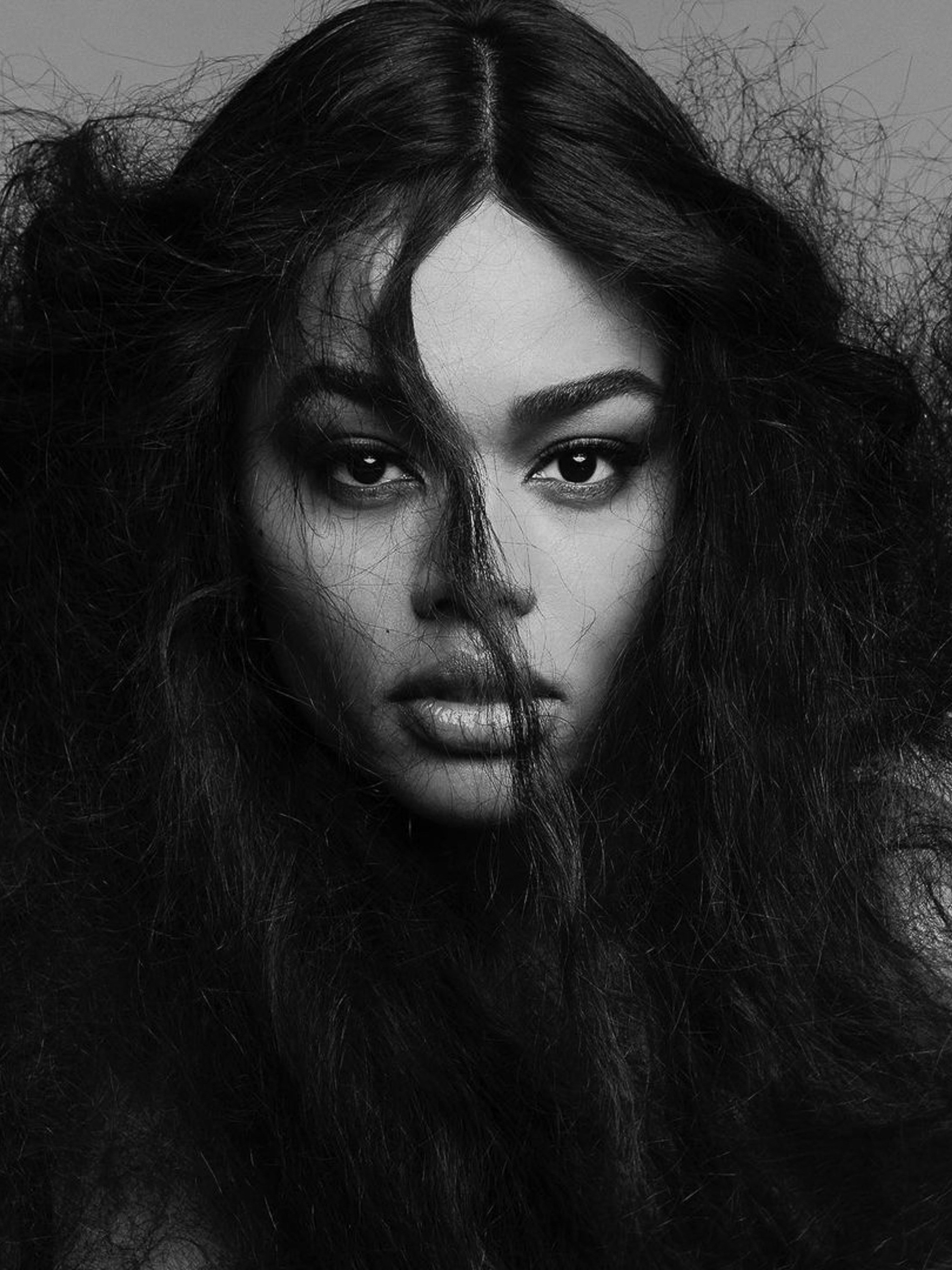 Monochrome portrait of an adult woman with long, dark wavy hair in a close-up headshot with a neutral facial expression in a studio.