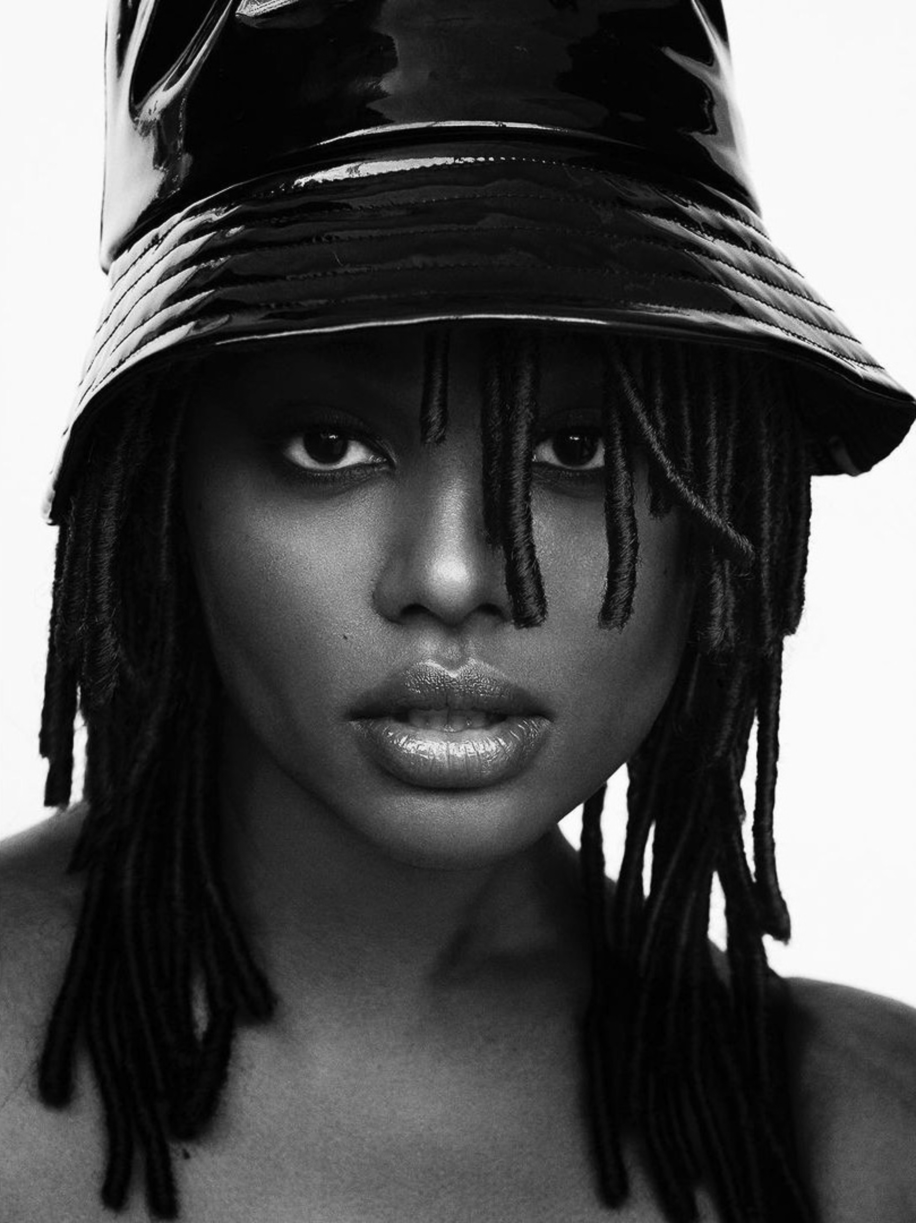 Close-up black and white portrait of a woman with dreadlocks wearing a glossy black bucket hat against a white background.