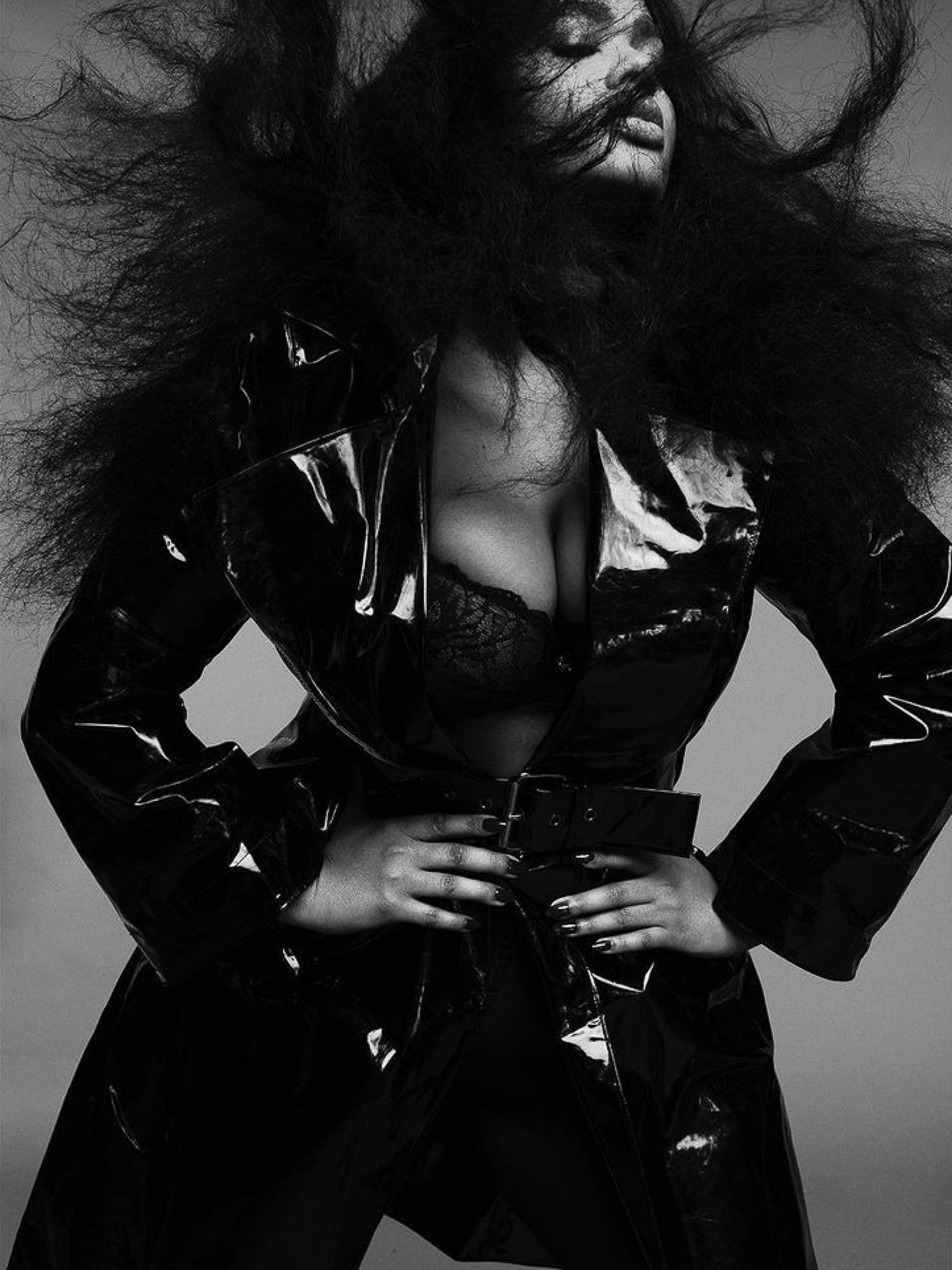 Black and white portrait of a woman with large afro wearing a shiny black trench coat and lace bra, posing with her hands on her waist.