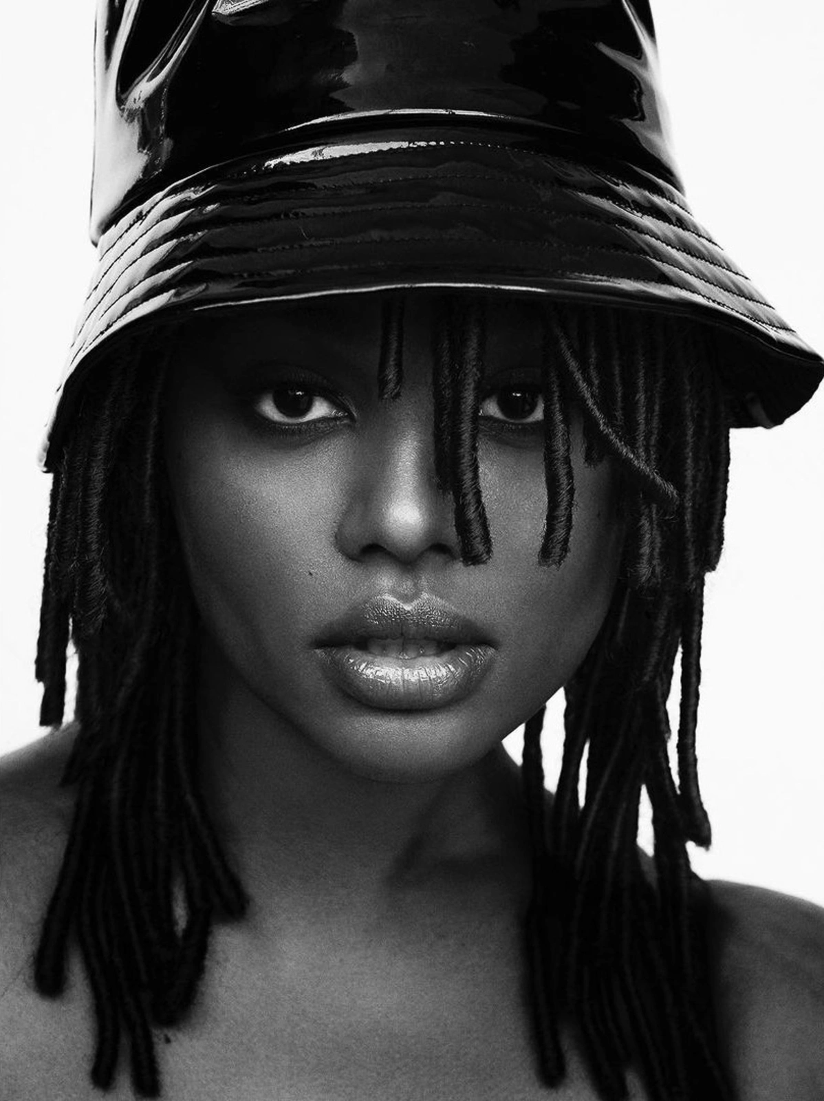 Close-up monochrome portrait of a Black woman wearing a glossy black bucket hat and locs for V Magazine editorial.