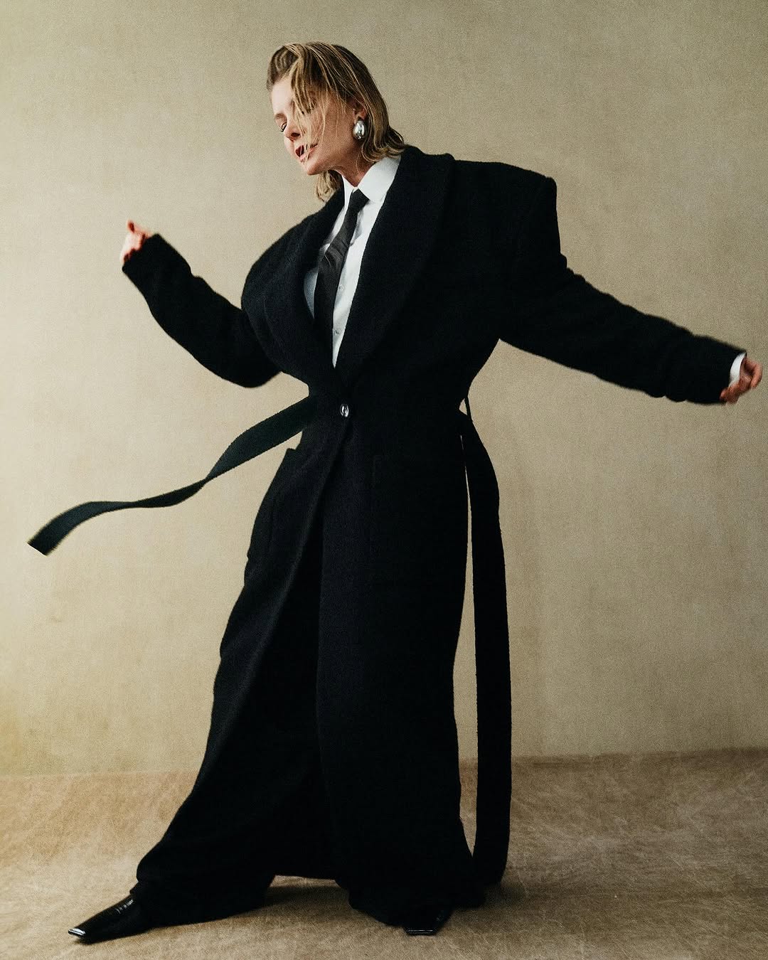 Elegant woman in an oversized black coat with a tie, posing in a light neutral-toned studio with soft lighting, and wearing pointed toe heels.