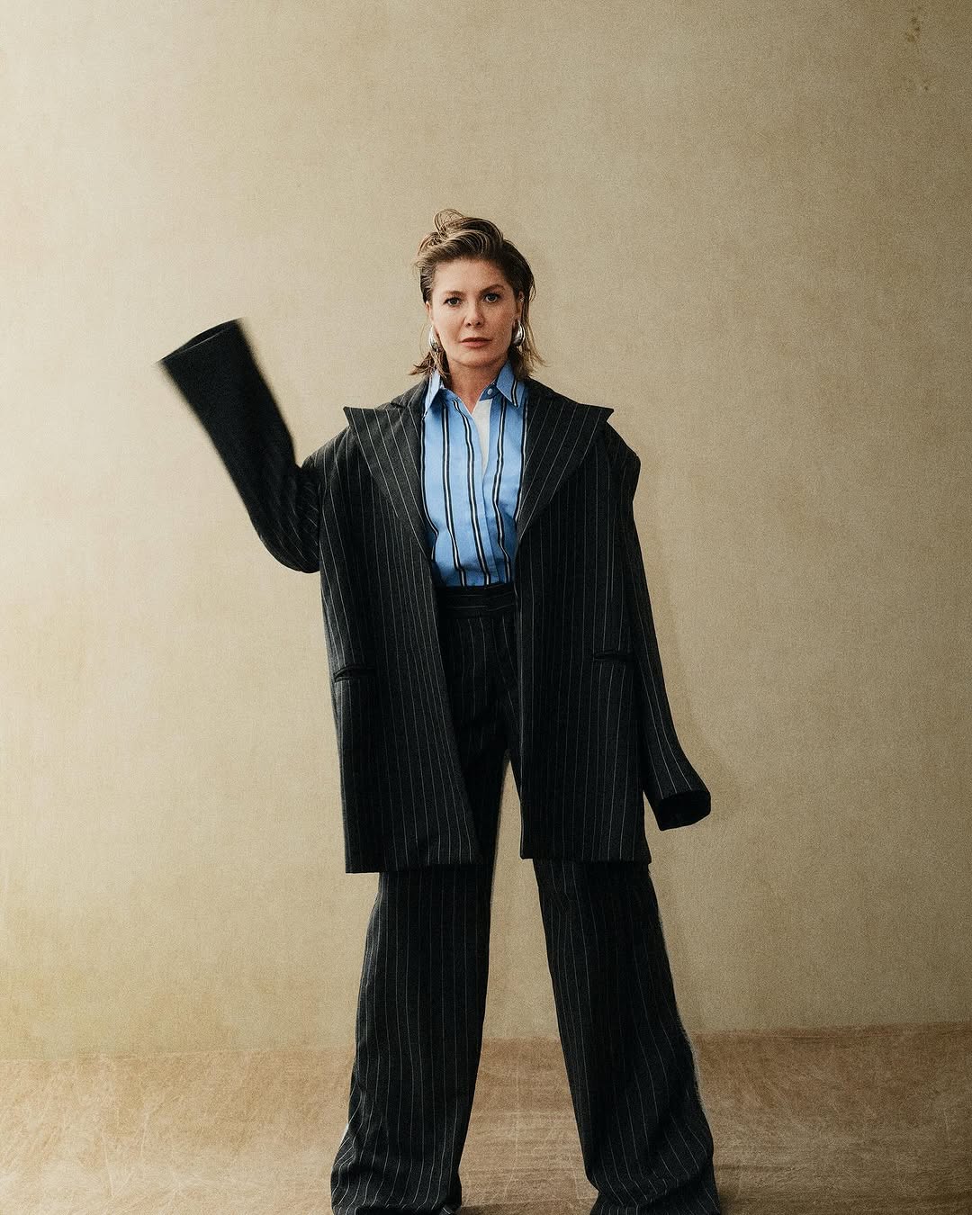 Fashion portrait of an adult woman wearing an oversized, striped suit with a light blue shirt, posing in a neutral-toned studio.