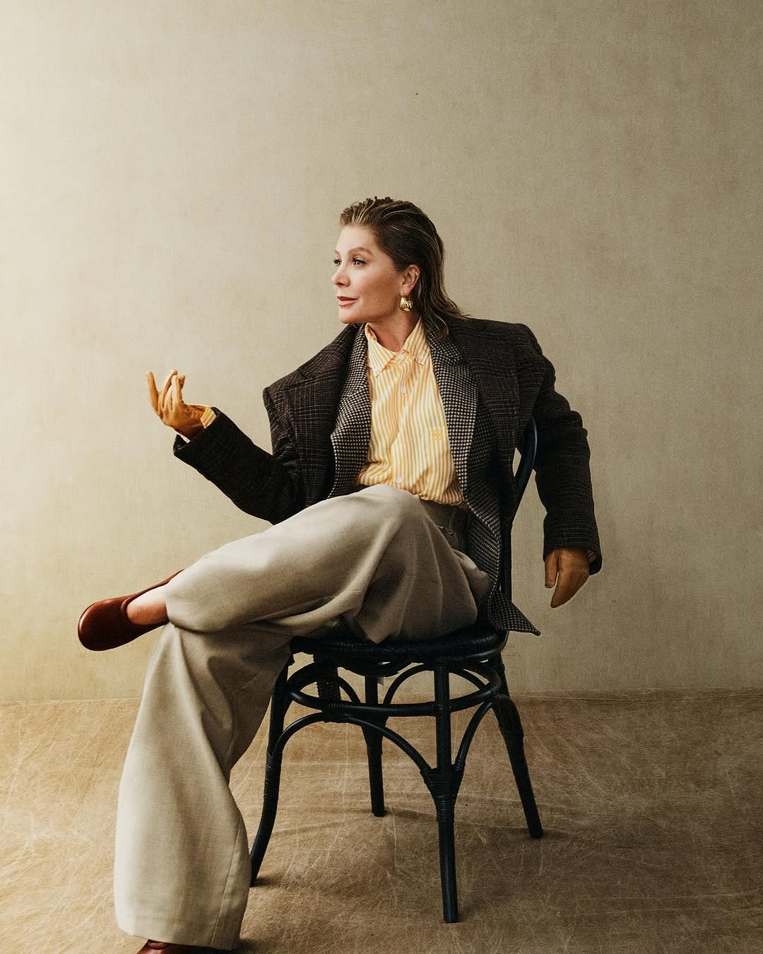 Elegant mature woman with wet hair, wearing a tweed blazer and wide-leg trousers, poses confidently on a black chair in a neutral studio.