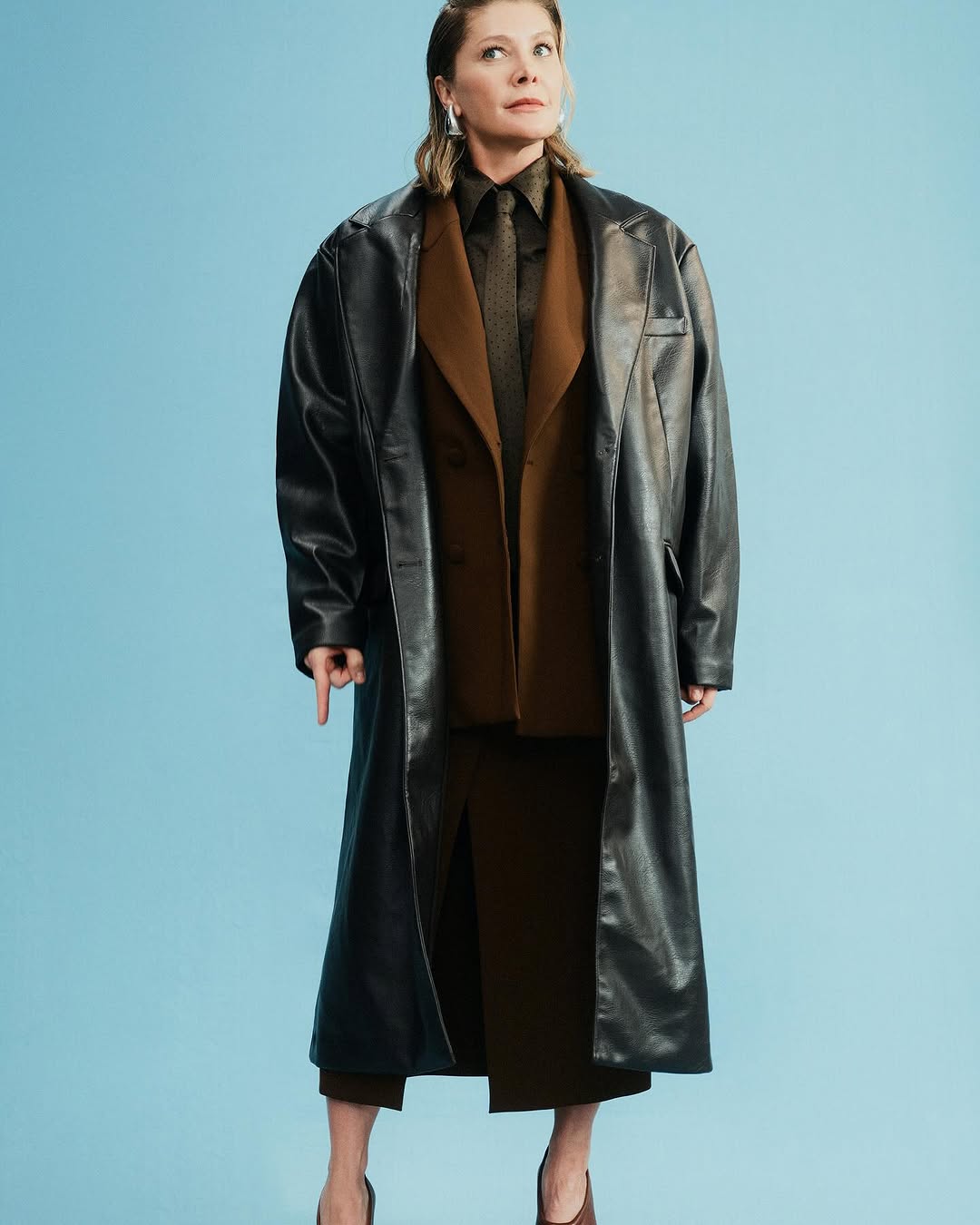 Studio shot of an adult woman in a black faux-leather trench coat, brown suit, and heels against a pale blue seamless background.