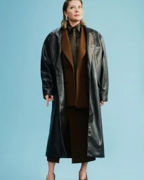 Natalie Bassingthwaighte wearing a long black leather coat over a brown suit and tie against a blue background