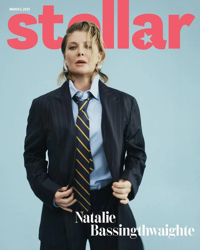 Woman wearing a dark pinstripe blazer, light blue shirt, and striped tie against a solid light blue background.
