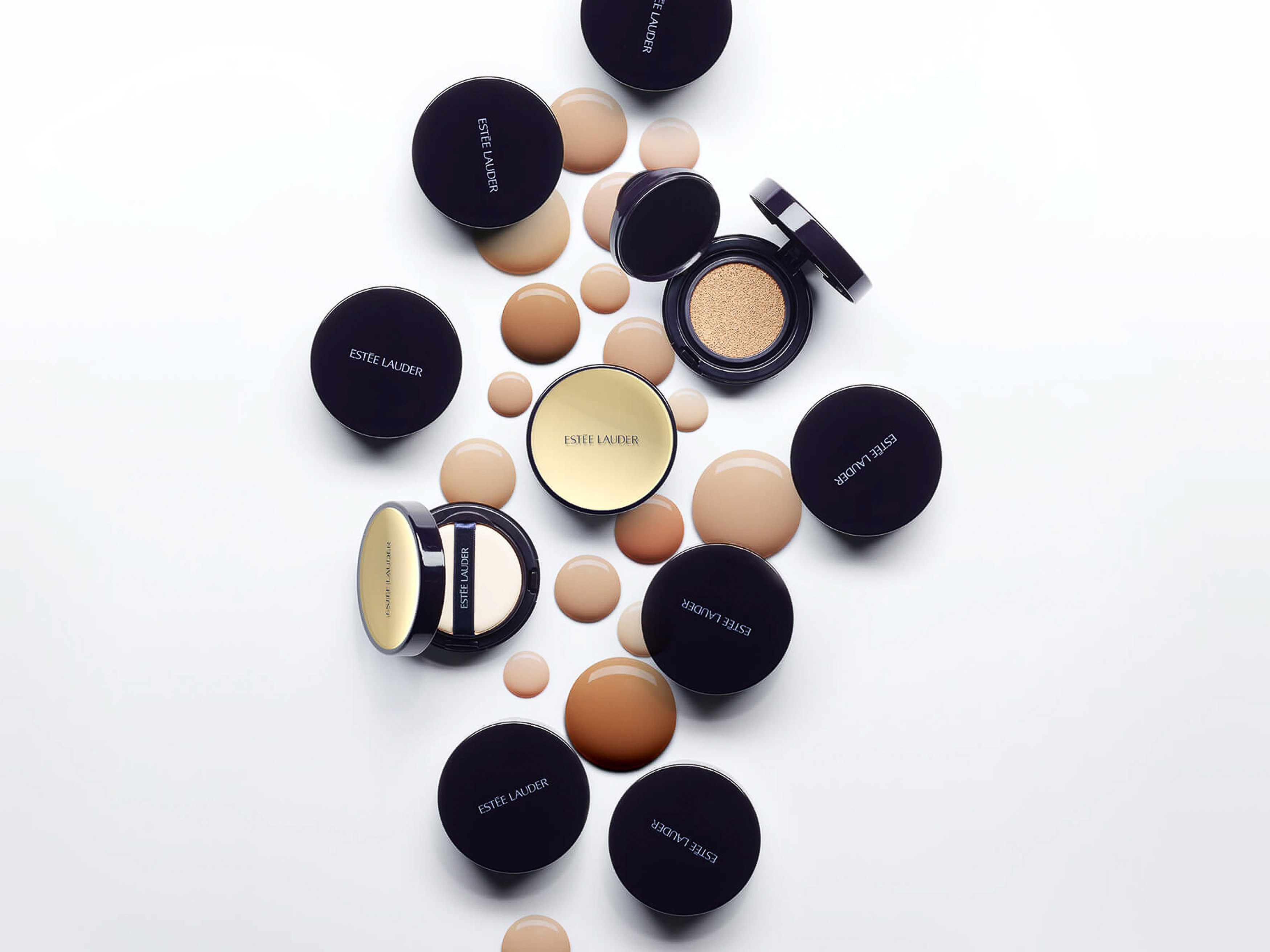 Flat lay studio shot of Estee Lauder makeup compacts and foundation droplets in tan and brown tones on a white surface.