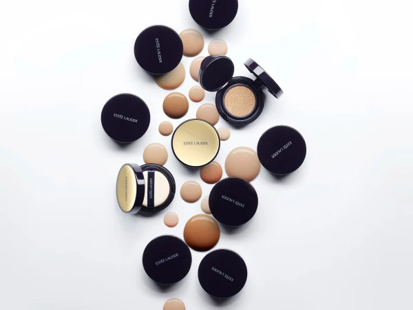 Flat lay studio shot of Estee Lauder makeup compacts and foundation droplets in tan and brown tones on a white surface.
