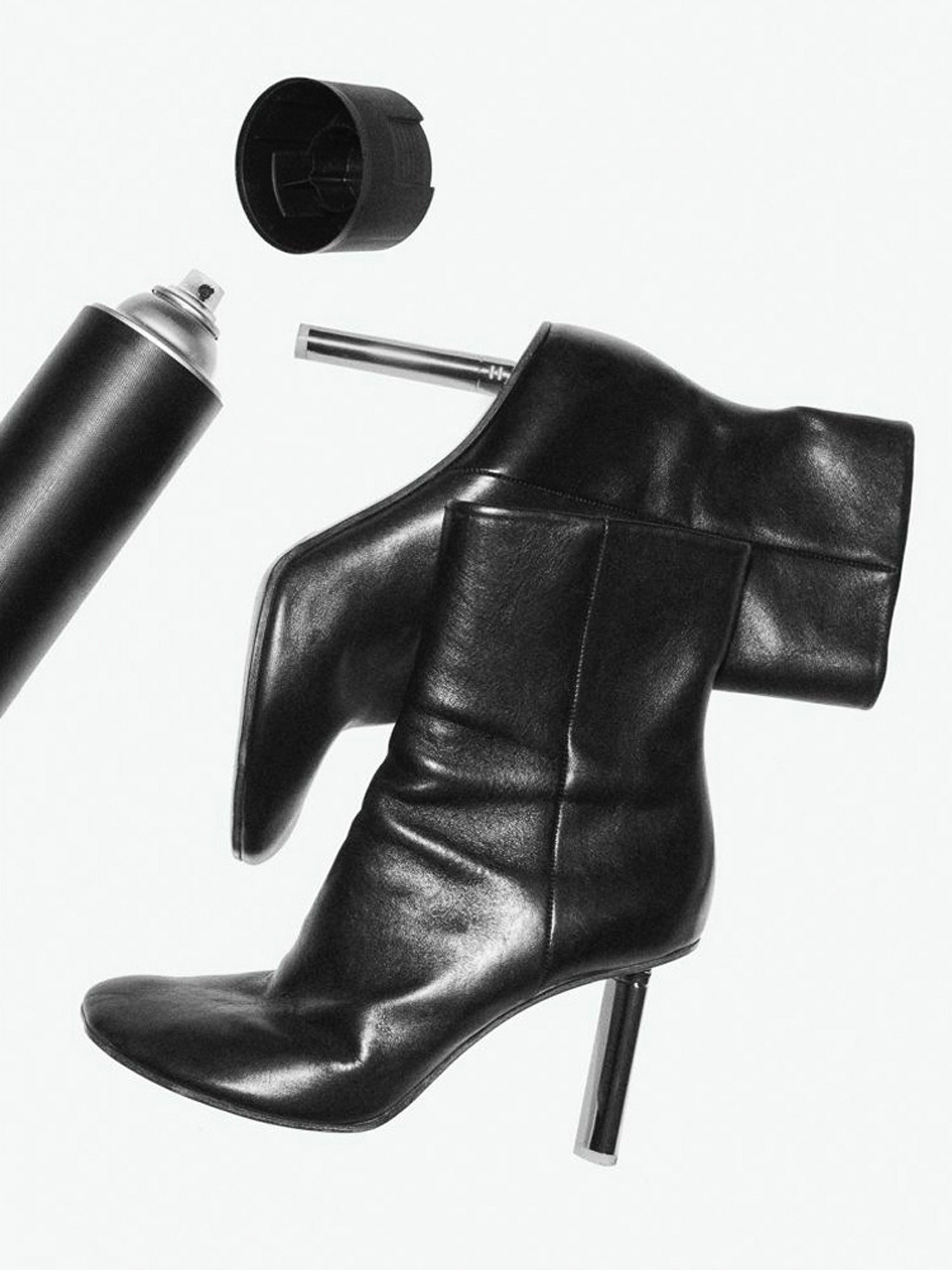 Stylized black and white still life photograph of shiny black high-heeled boots with a spray paint can and cap on a white backdrop.