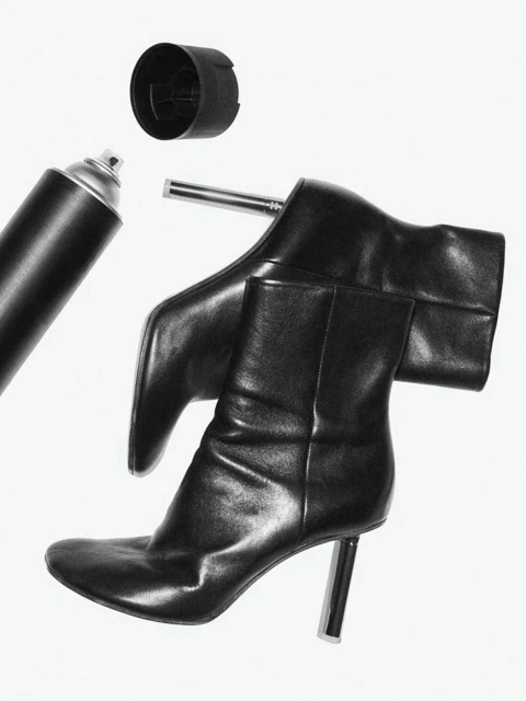 Stylized black and white still life photograph of shiny black high-heeled boots with a spray paint can and cap on a white backdrop.