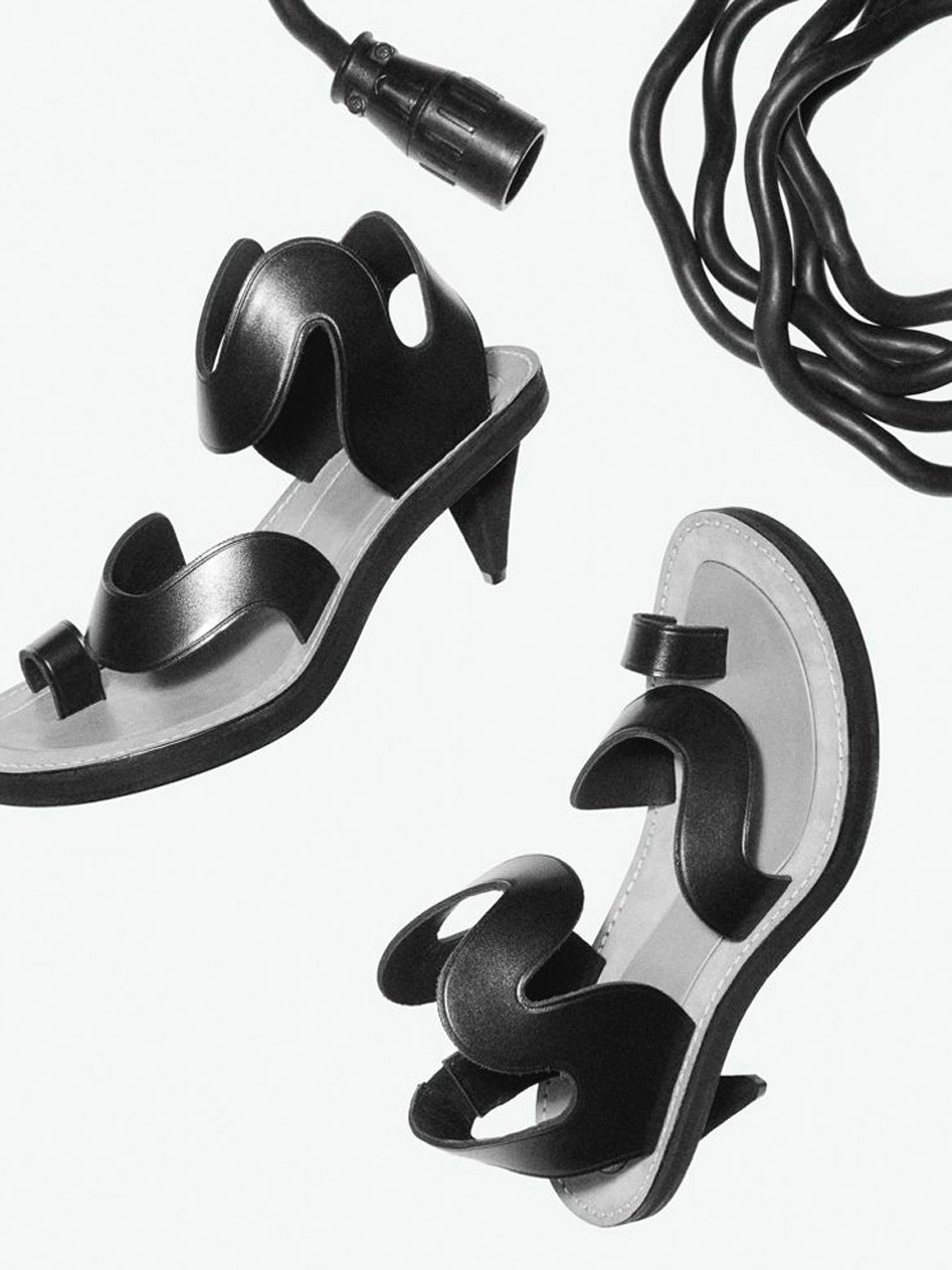 Still life of two black heeled sandals, with toe loops and wavy straps, arranged beside black electrical cable on a white background.