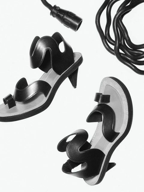 Still life of two black heeled sandals, with toe loops and wavy straps, arranged beside black electrical cable on a white background.