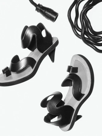 Still life of two black heeled sandals, with toe loops and wavy straps, arranged beside black electrical cable on a white background.