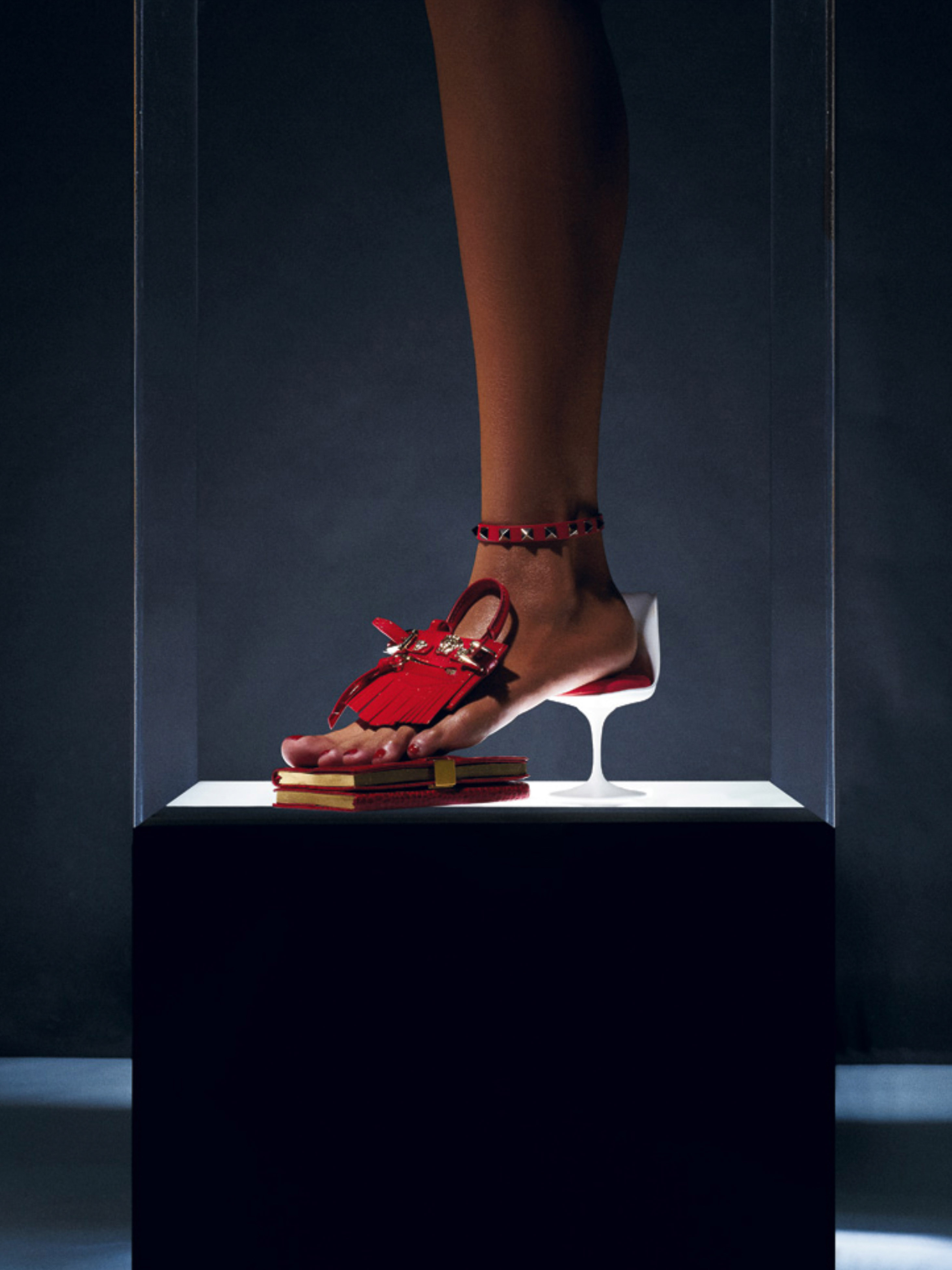 Foot in red sandal with ankle bracelet steps on books in clear display case, fashion-forward and visually striking. 