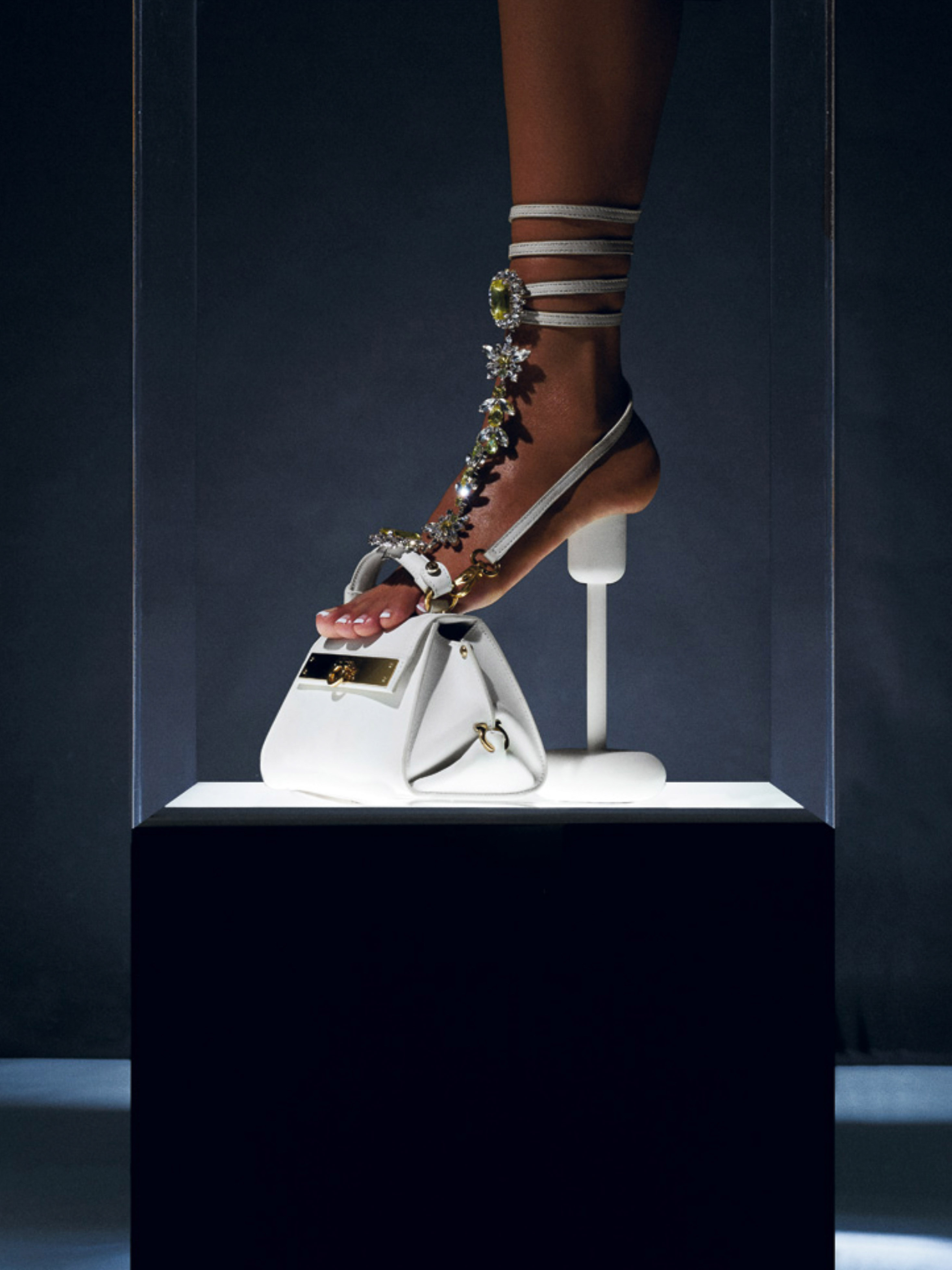Foot with designer embellished heel and ankle strap, standing on a white leather handbag displayed in a museum-like clear display case.