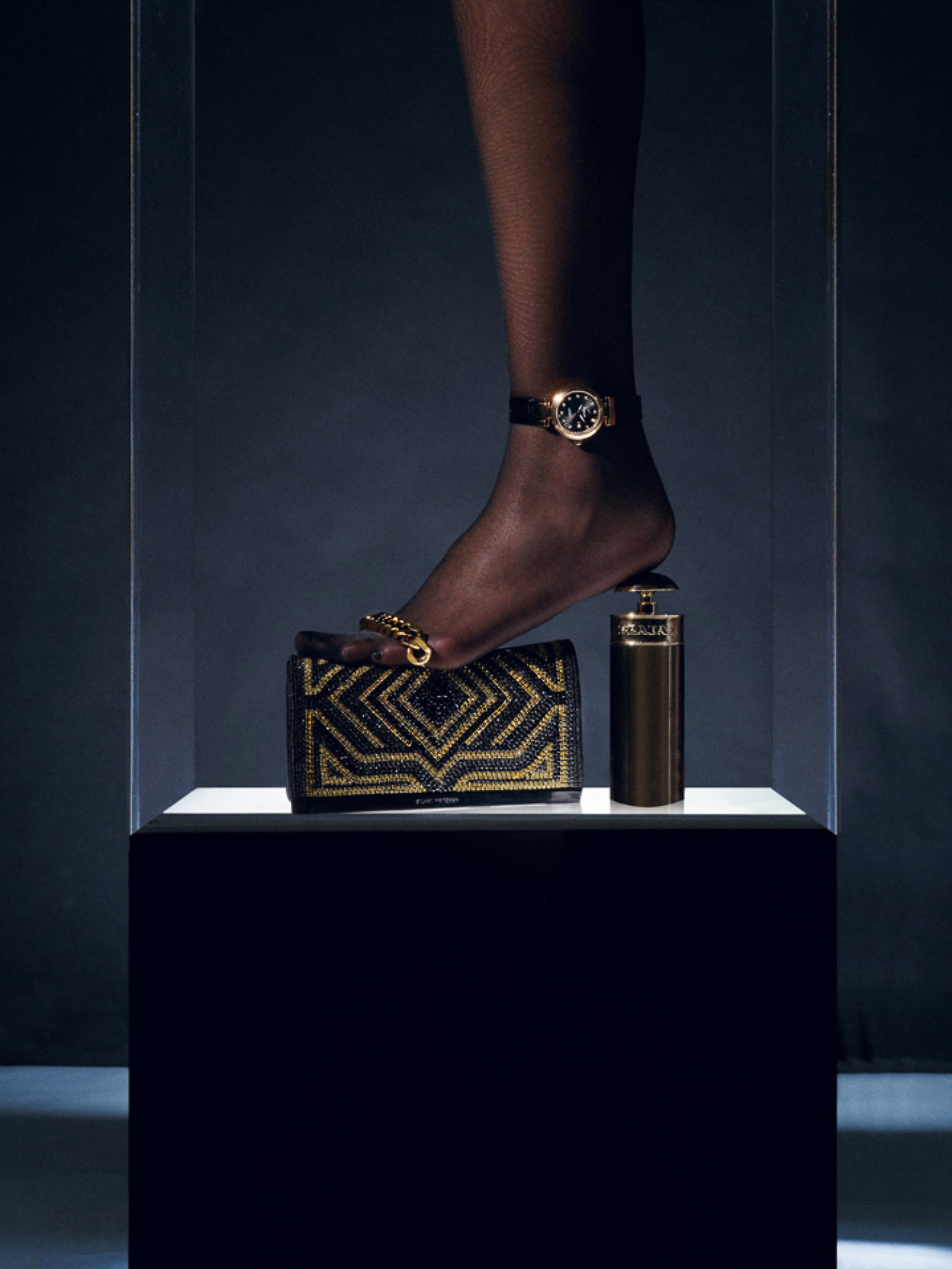 A black woman's foot with jewelry and a wristwatch steps on a black and gold handbag and perfume bottle on a white pedestal.