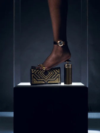 A black woman's foot with jewelry and a wristwatch steps on a black and gold handbag and perfume bottle on a white pedestal.